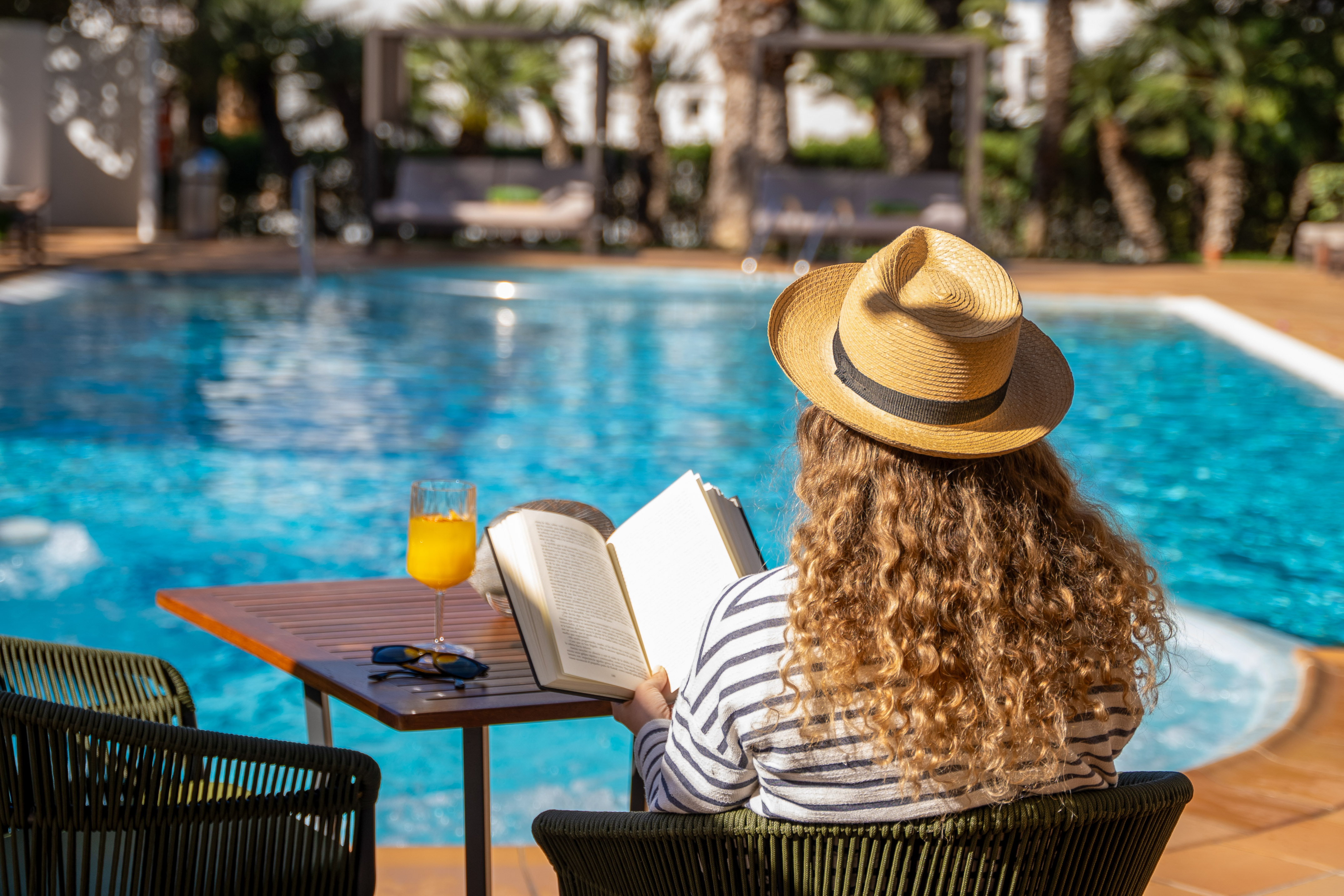 Woman in straw hat reading book by sunny resort pool with a drink.