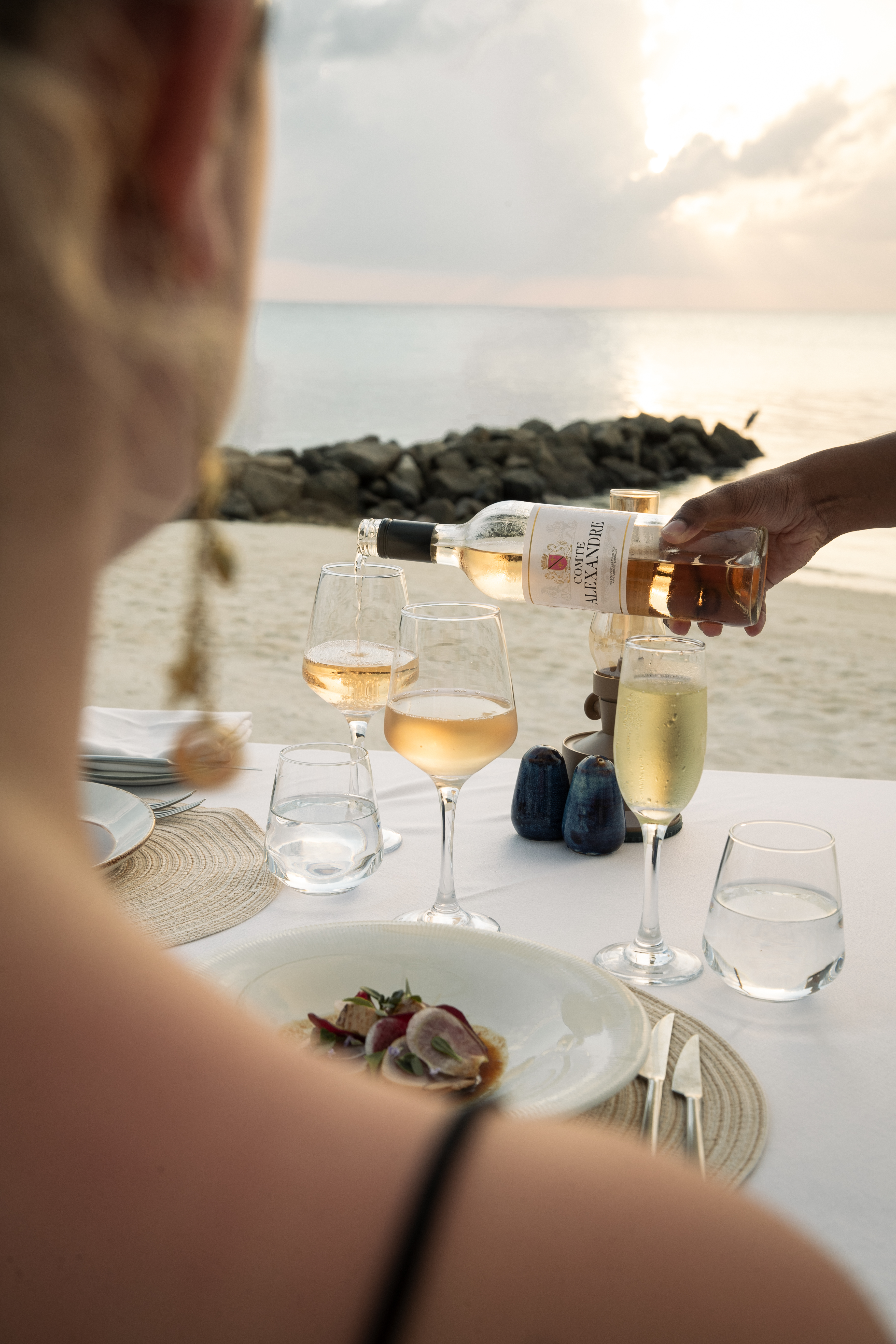 Hand pours rosé wine at a romantic beach dinner with gourmet food, sunset.
