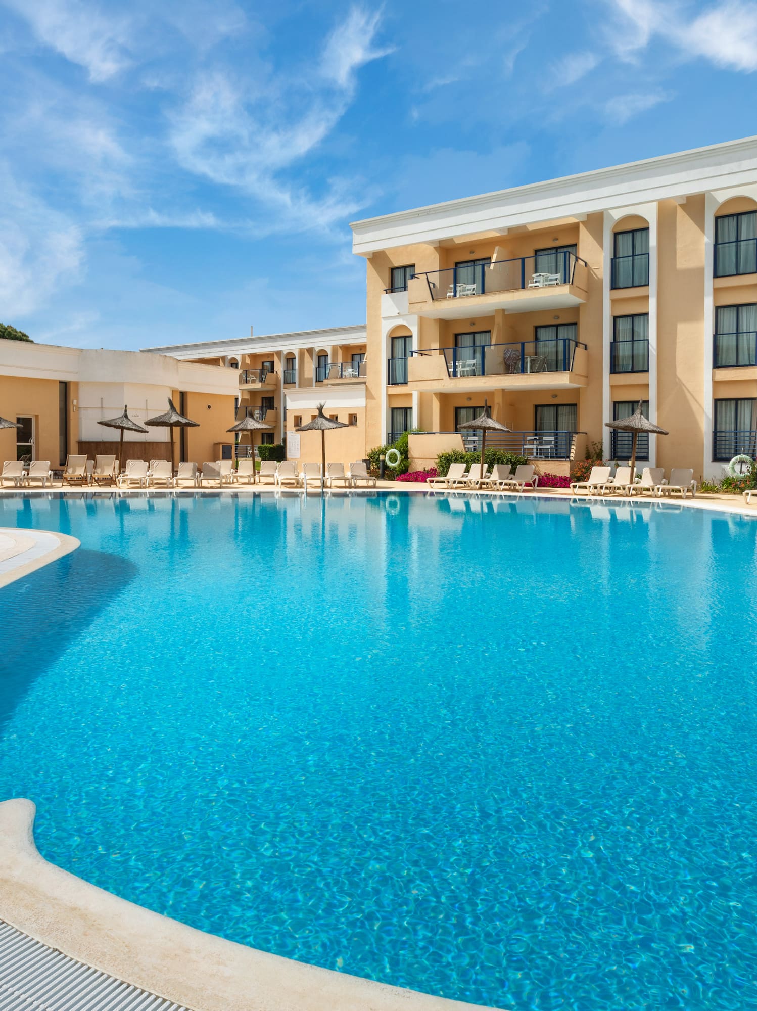 Sunny hotel pool area with lounge chairs, palm tree, and resort building.