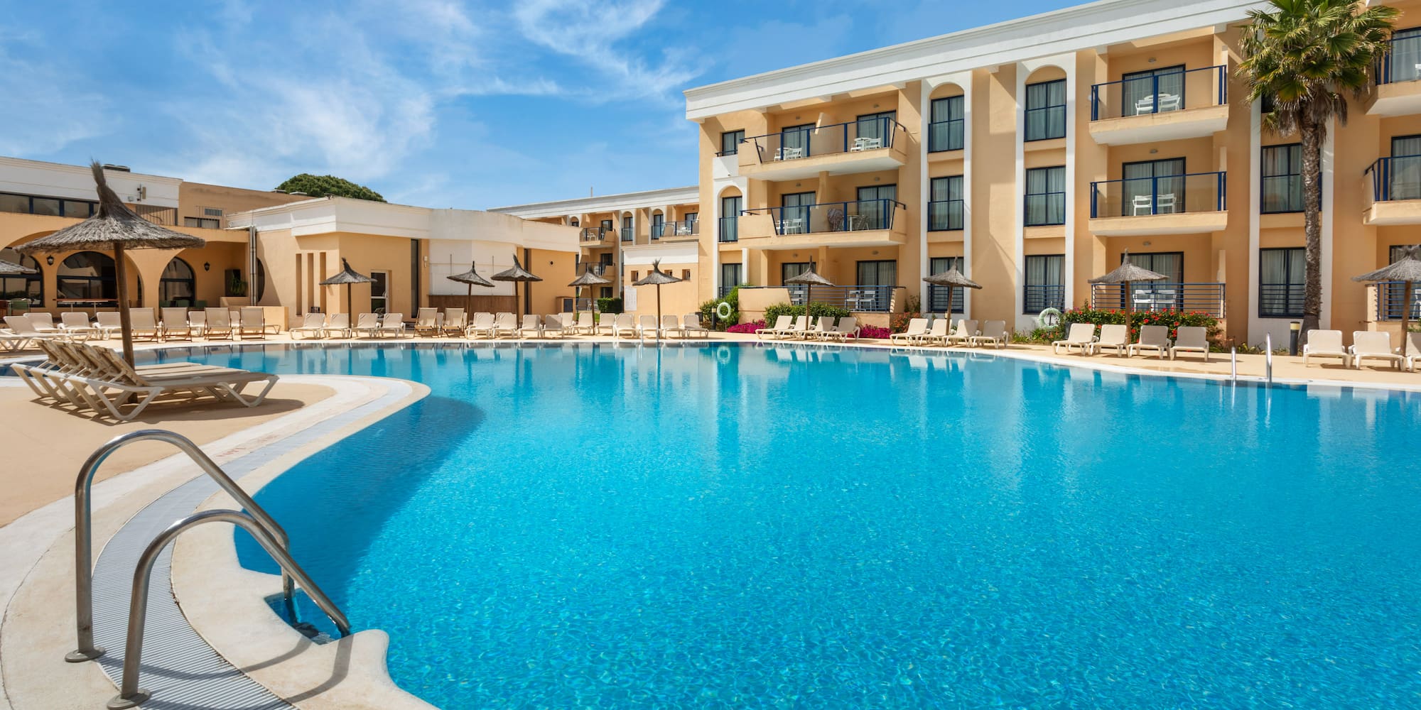 Hotel resort swimming pool with sun loungers, balconies, and blue sky.