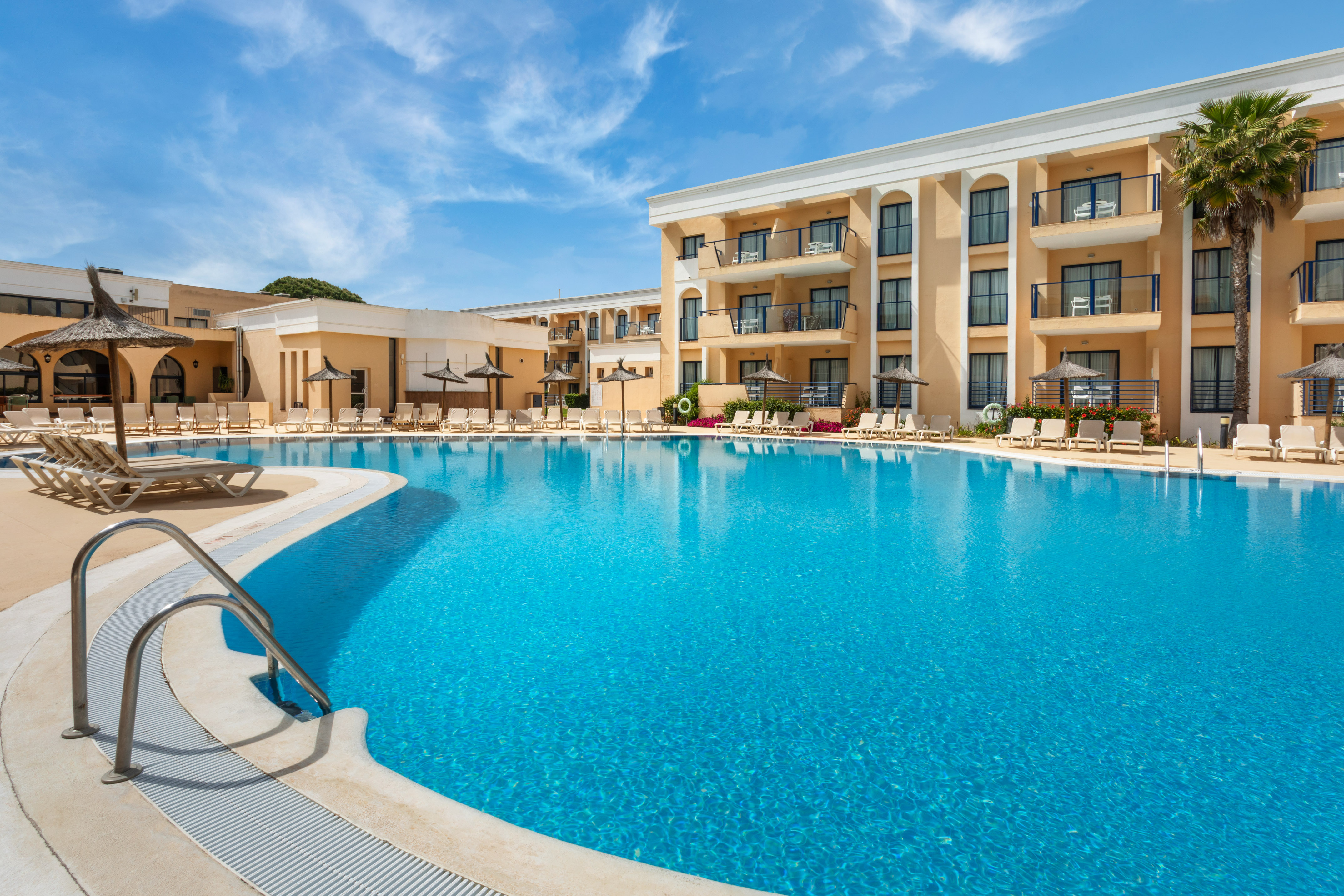 Hotel resort swimming pool with sun loungers, balconies, and blue sky.