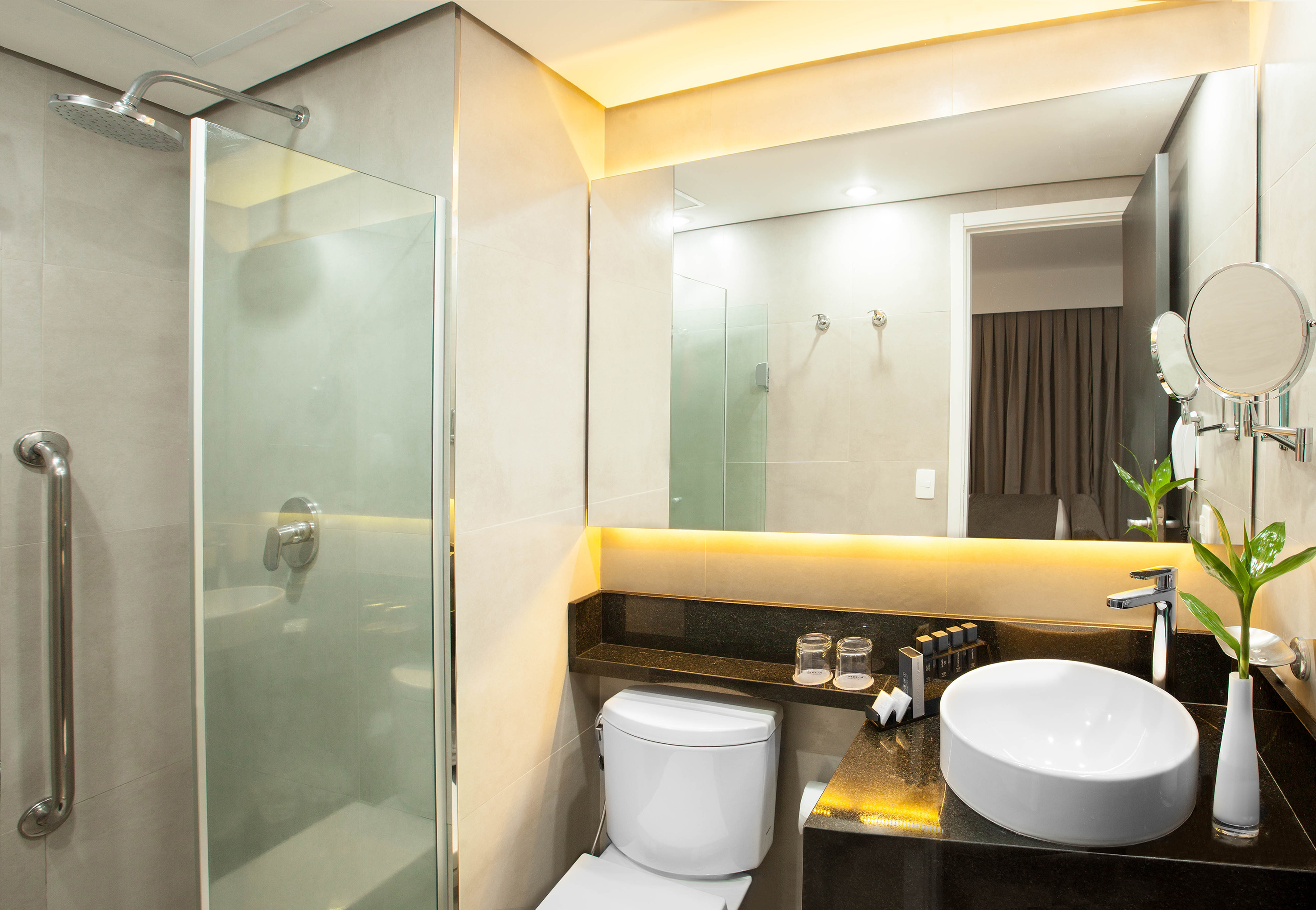 Modern hotel bathroom with glass shower, toilet, vessel sink, and backlit mirror.