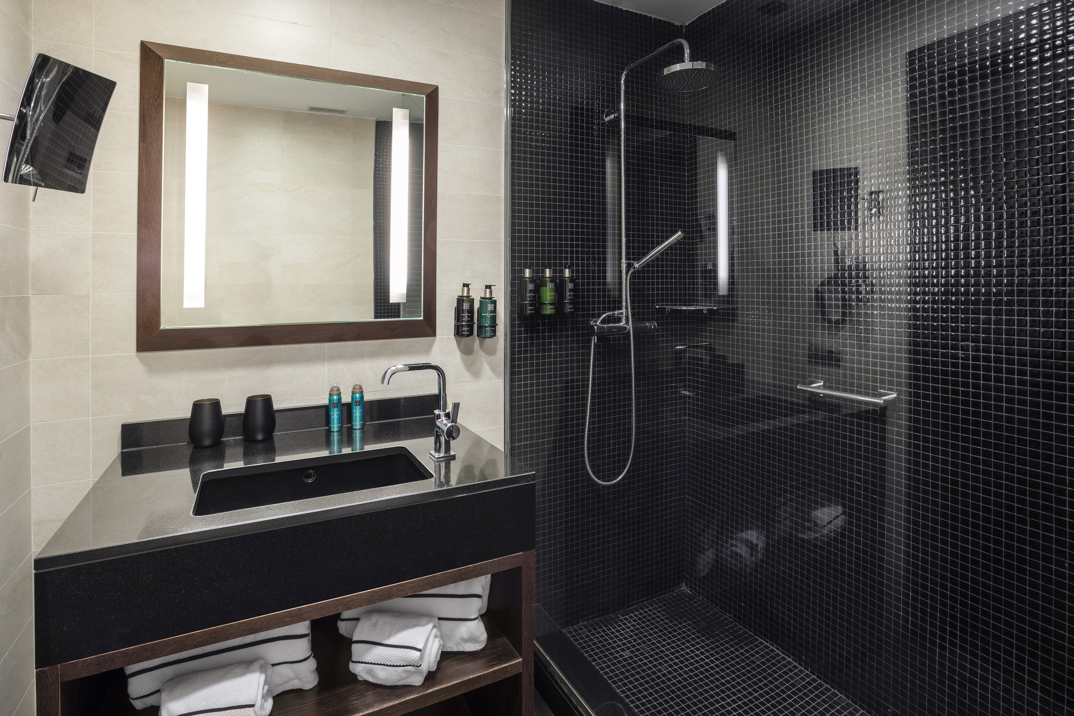 Stylish hotel bathroom with black shower, dark vanity, and lit mirror.