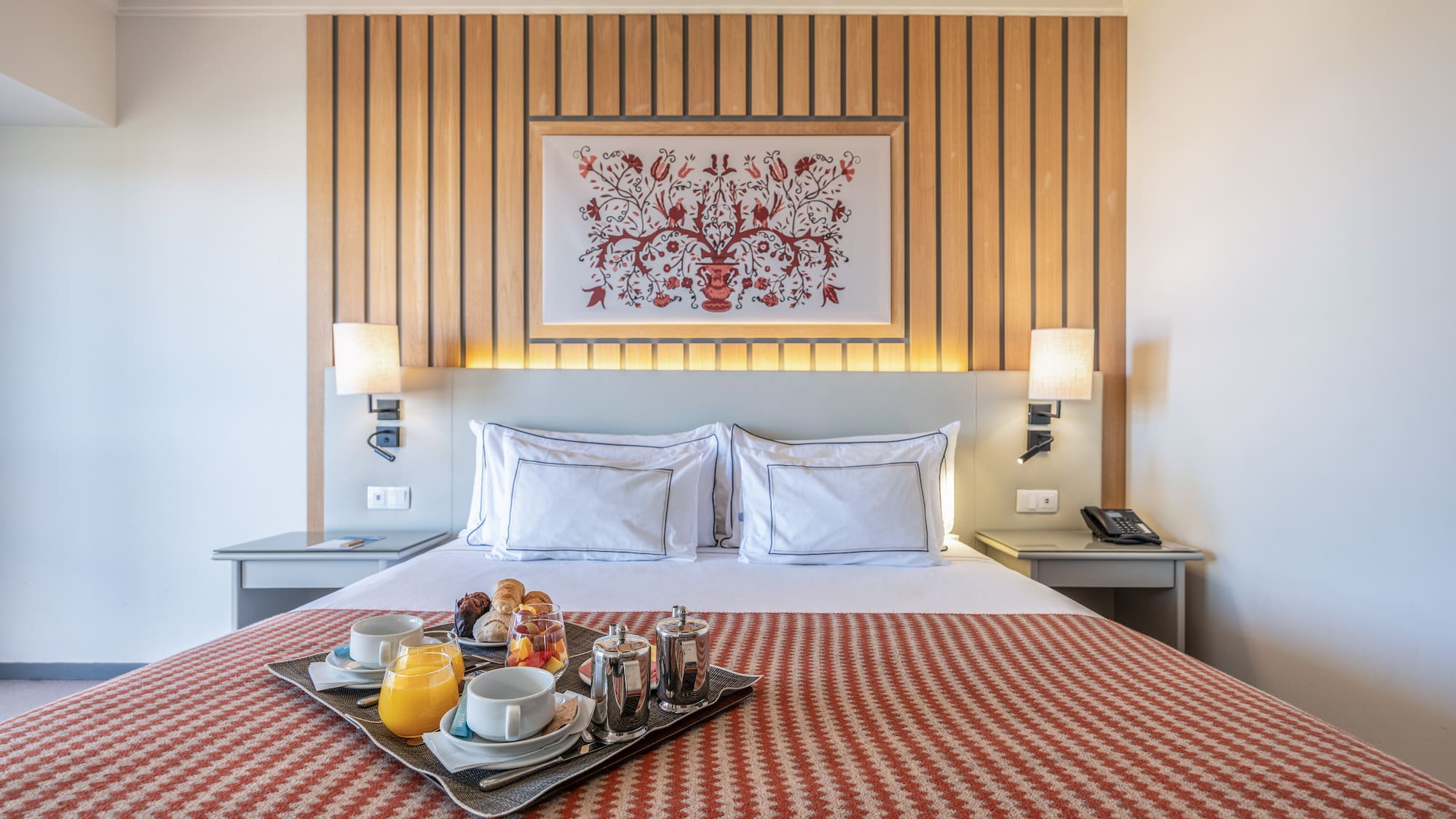 Hotel room with breakfast in bed, wooden headboard, and patterned bedspread.