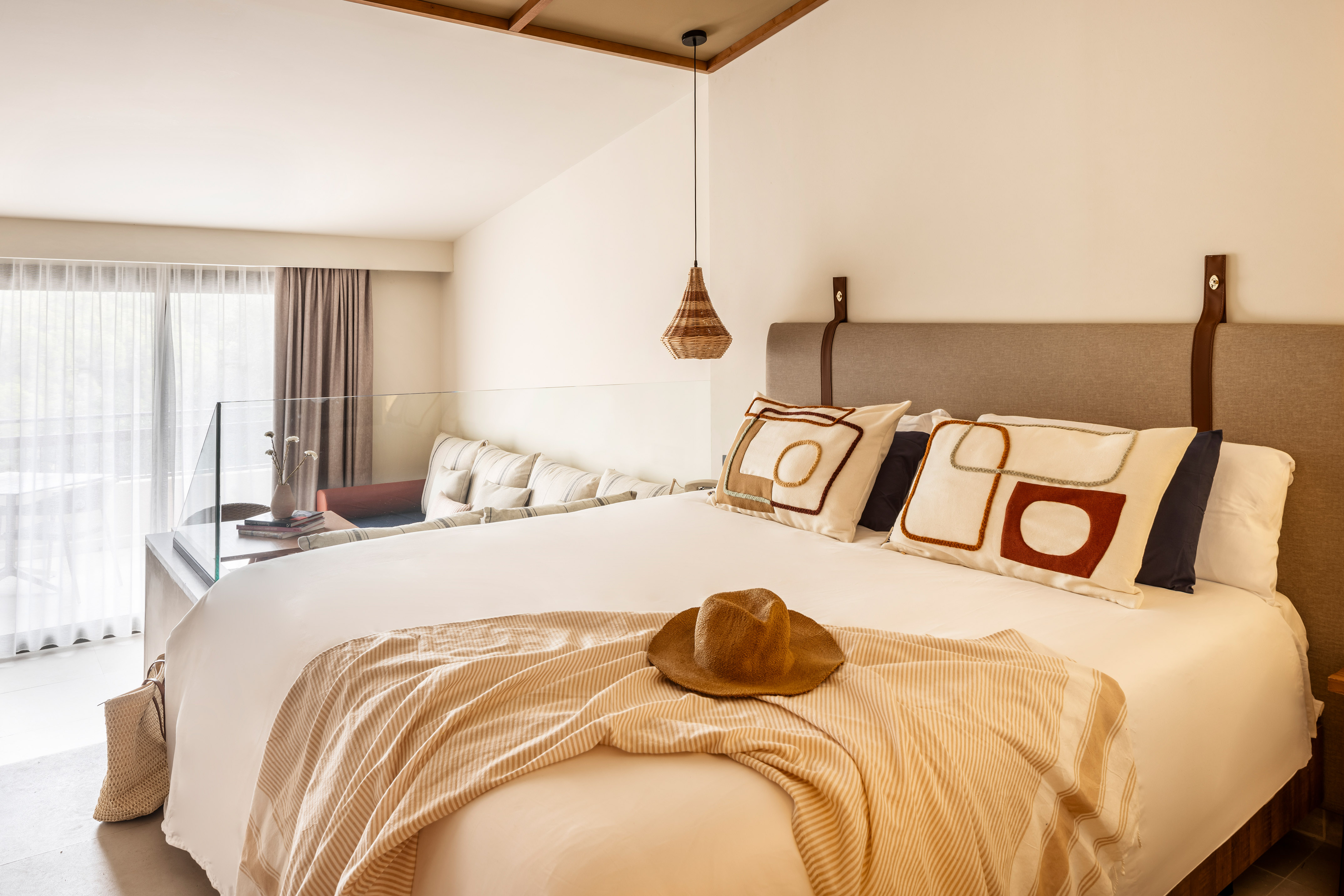 Modern hotel bedroom with large bed, straw hat, and separate lounge area.