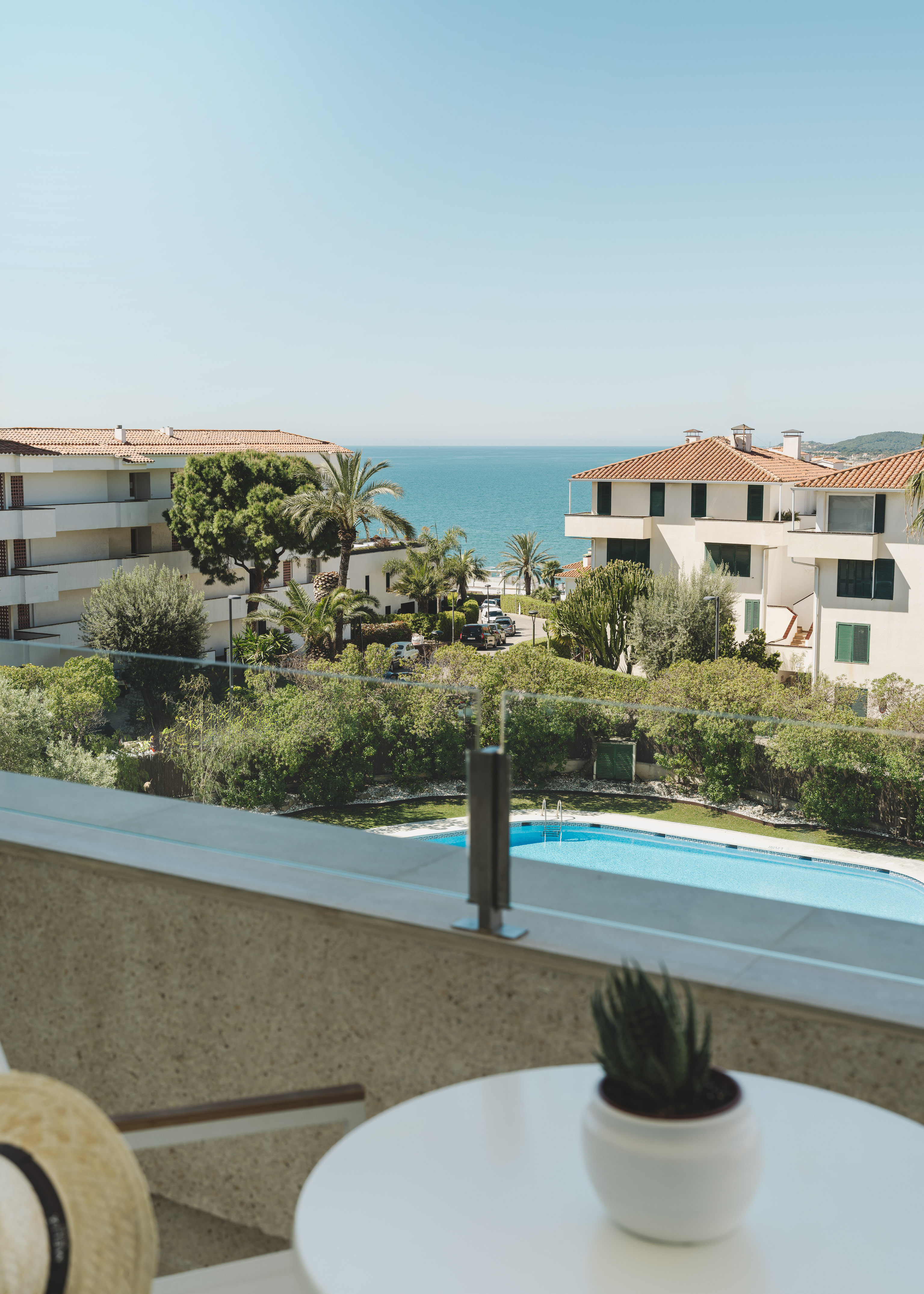 Balcony view of coastal resort with swimming pool, buildings, palm trees, and blue sea.