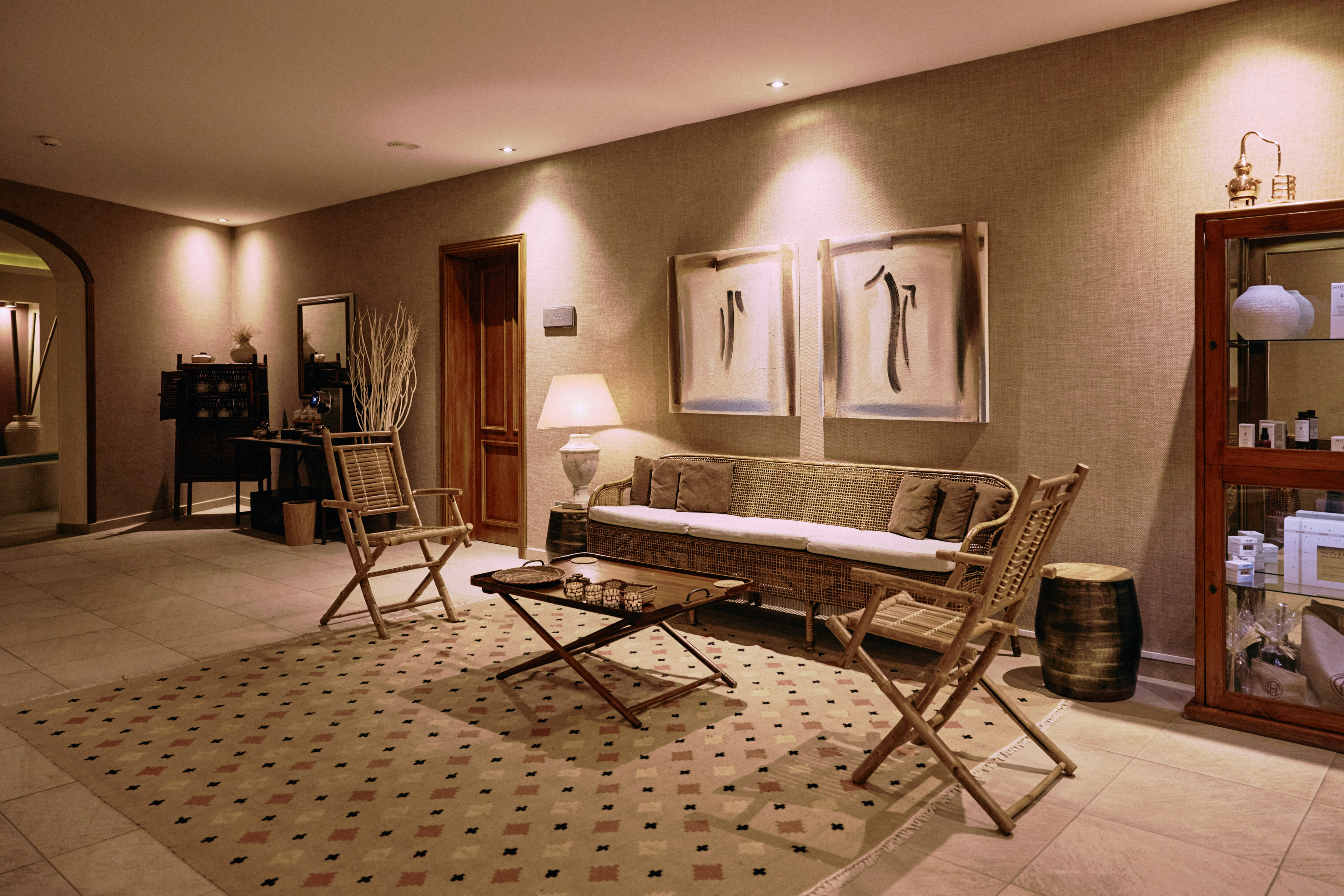Relaxing spa lounge with rattan sofa, wooden chairs, abstract art, and warm lighting.