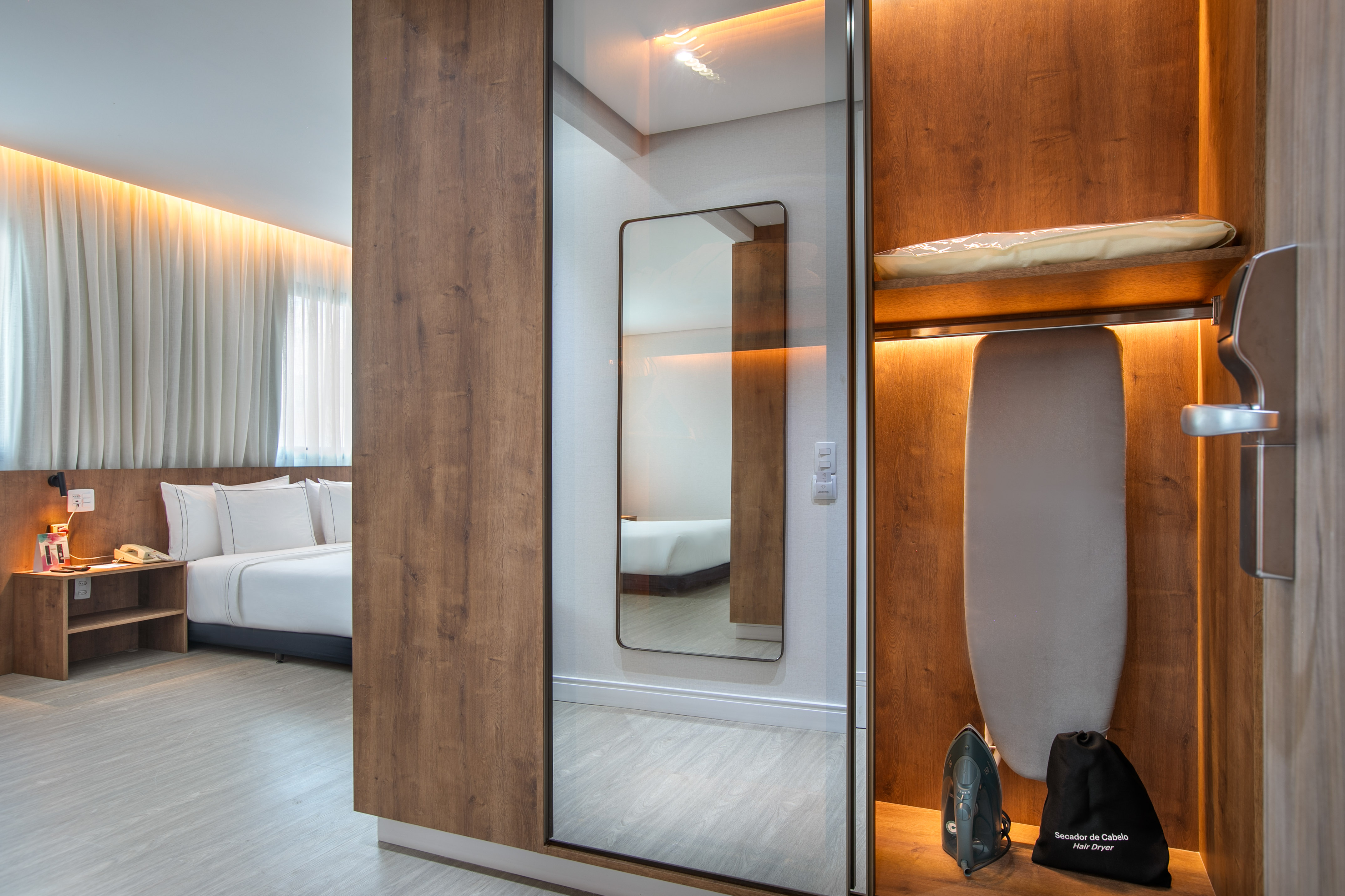 Modern hotel room: bed, wooden closet with mirror, ironing board, hairdryer.