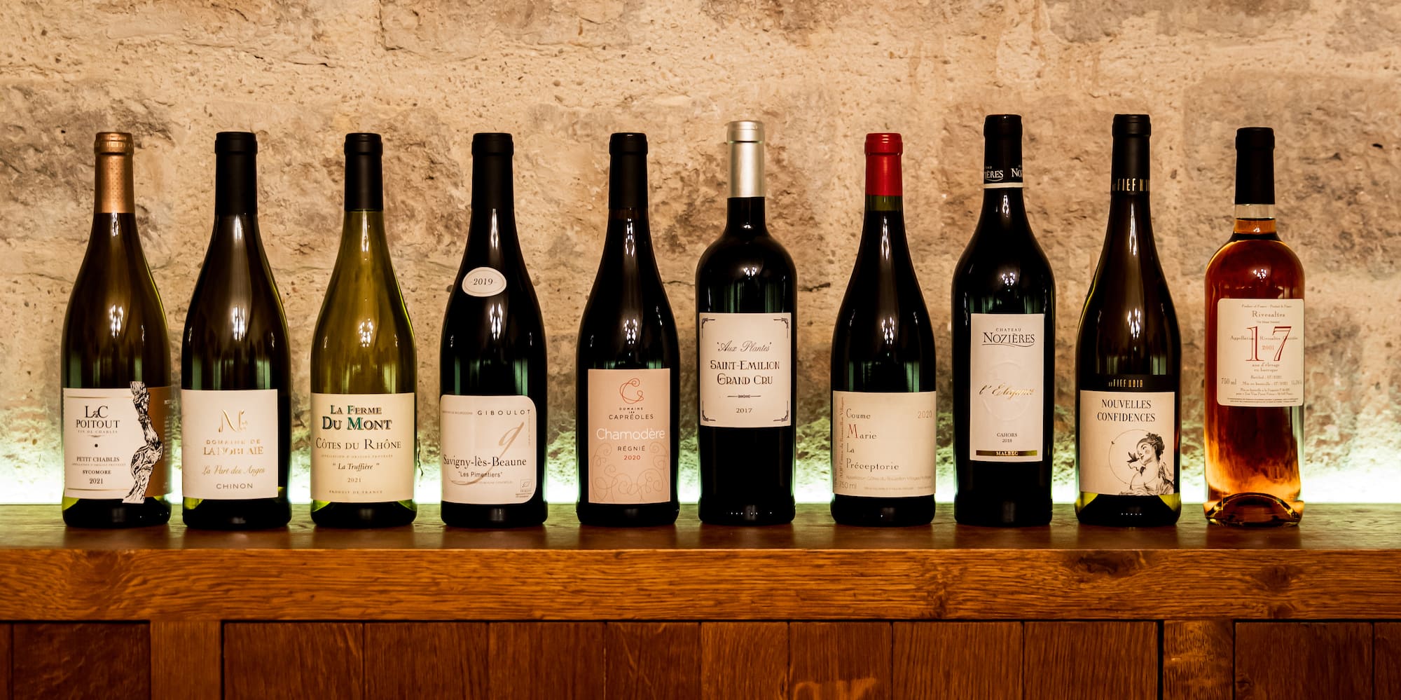 Ten diverse wine bottles on a lit wooden shelf against a rustic stone wall.