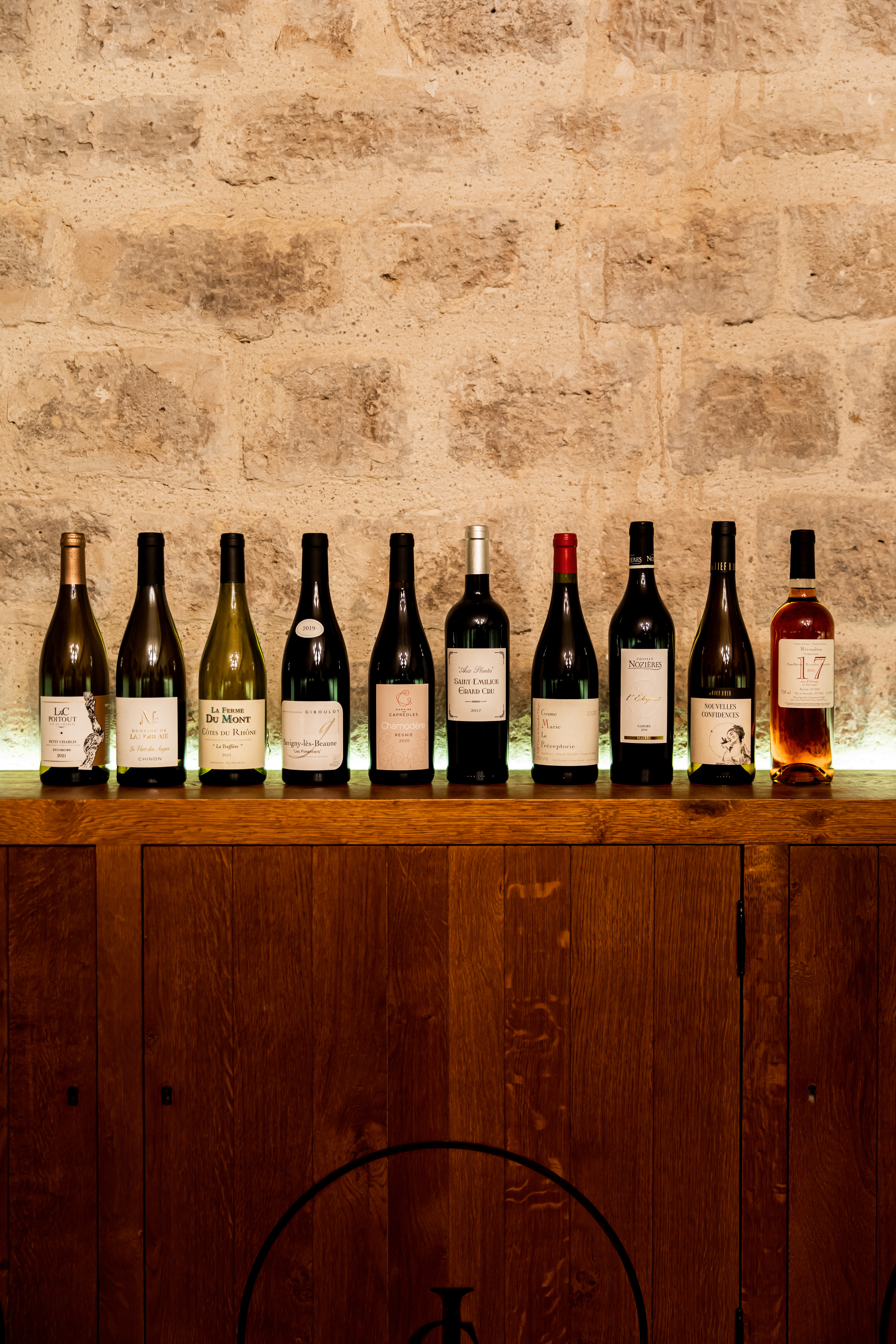 Ten diverse wine bottles on a lit wooden shelf against a rustic stone wall.