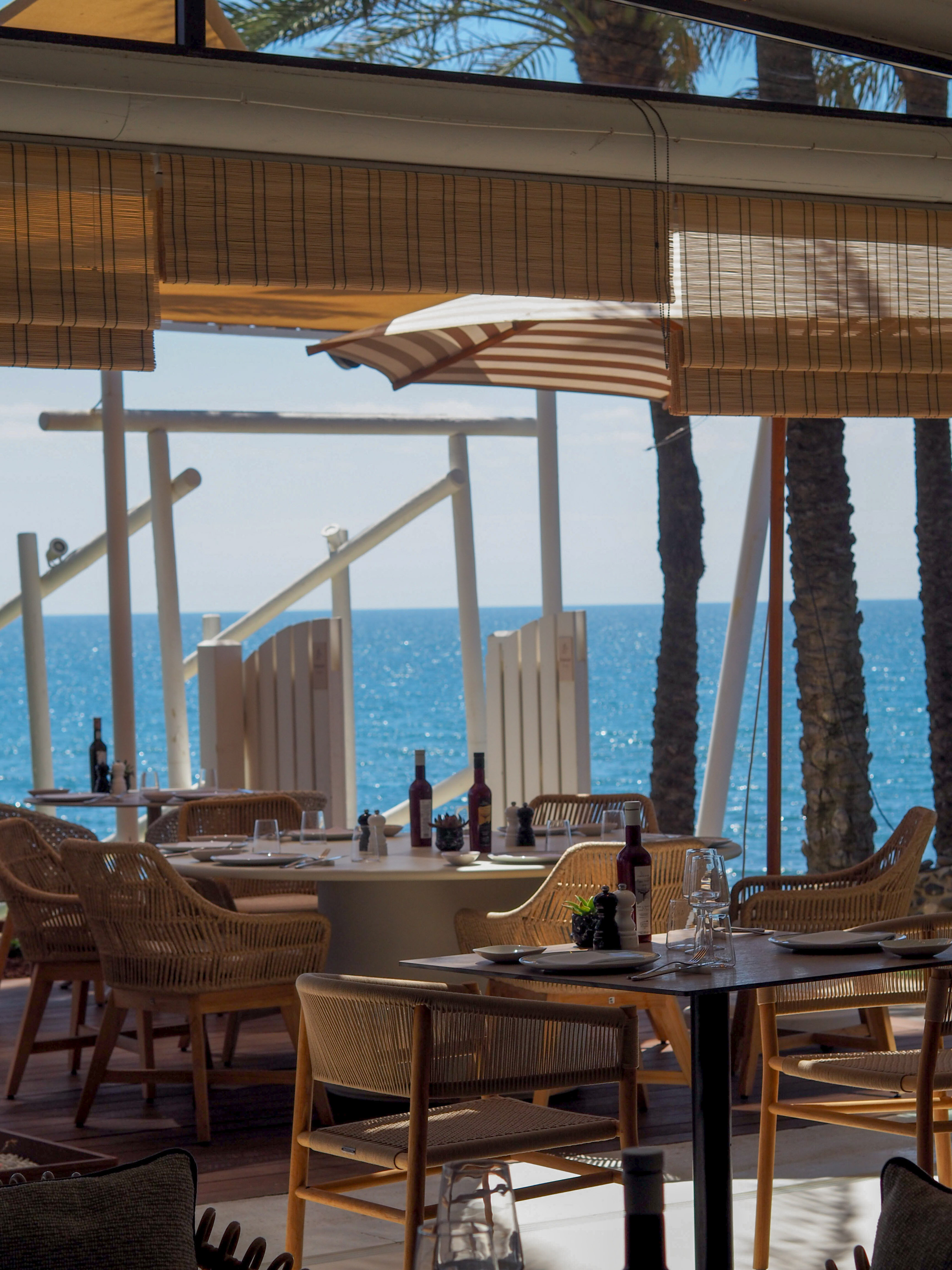 Seaside restaurant with set tables, rattan chairs, and sparkling blue ocean.