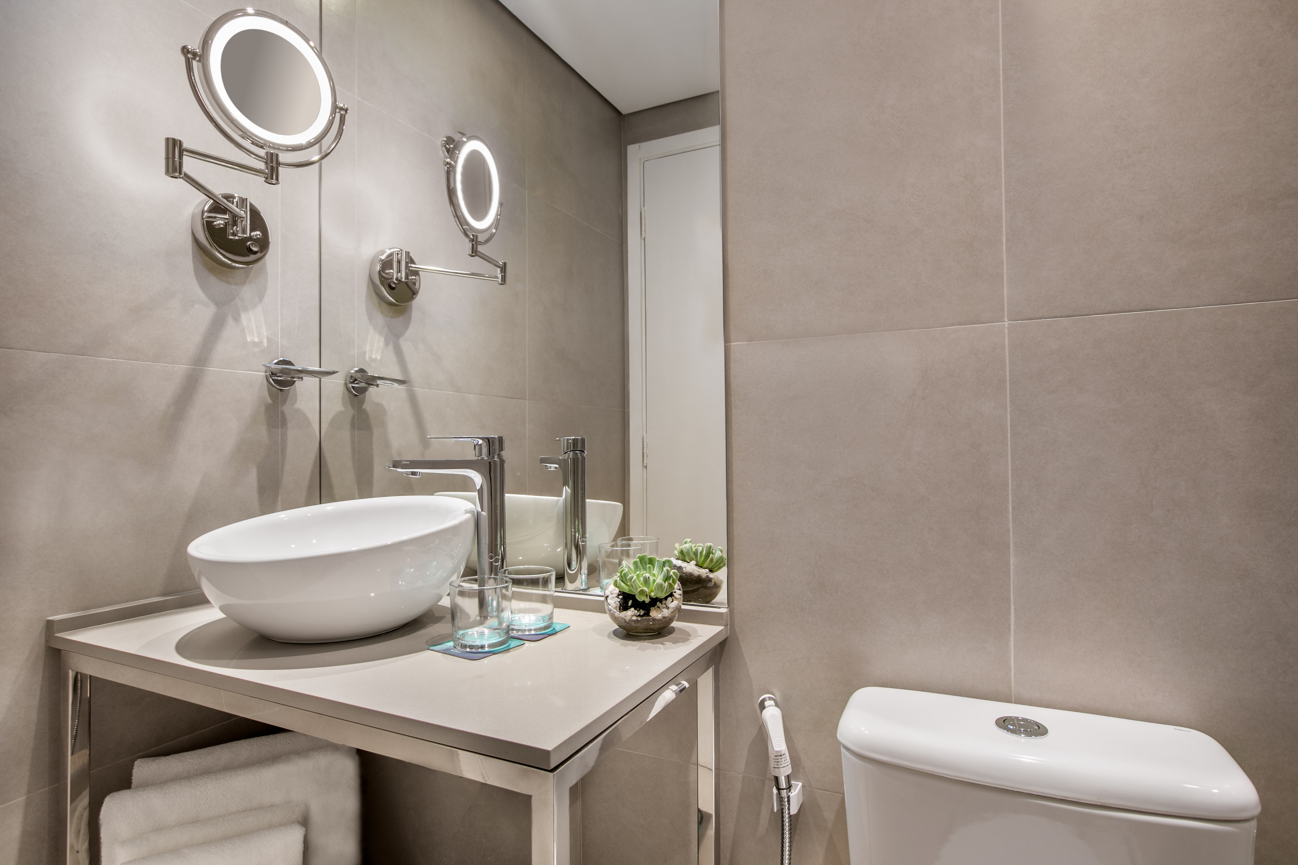 Contemporary hotel bathroom with vessel sink, lighted mirrors, and tiled walls.