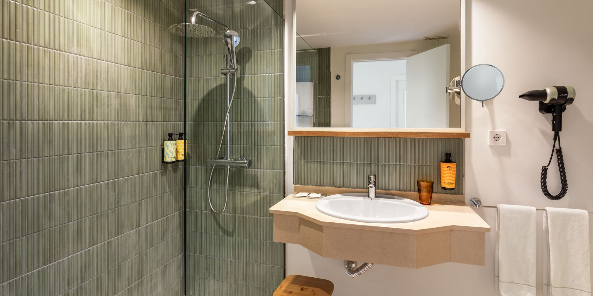 Modern hotel bathroom with green tiled shower, rain head, vanity, sink, and mirror.