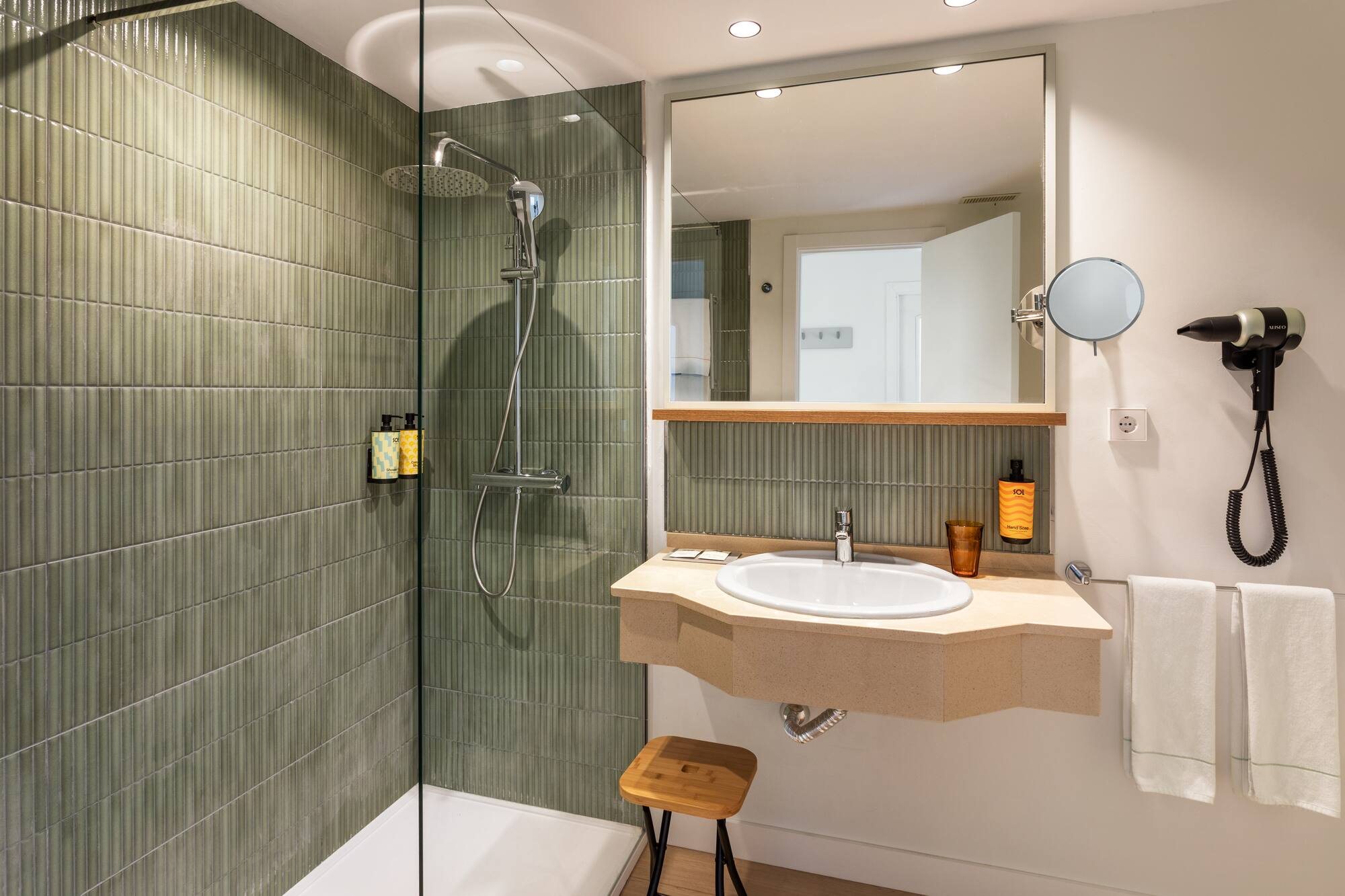 Modern hotel bathroom with green tiled shower, rain head, vanity, sink, and mirror.