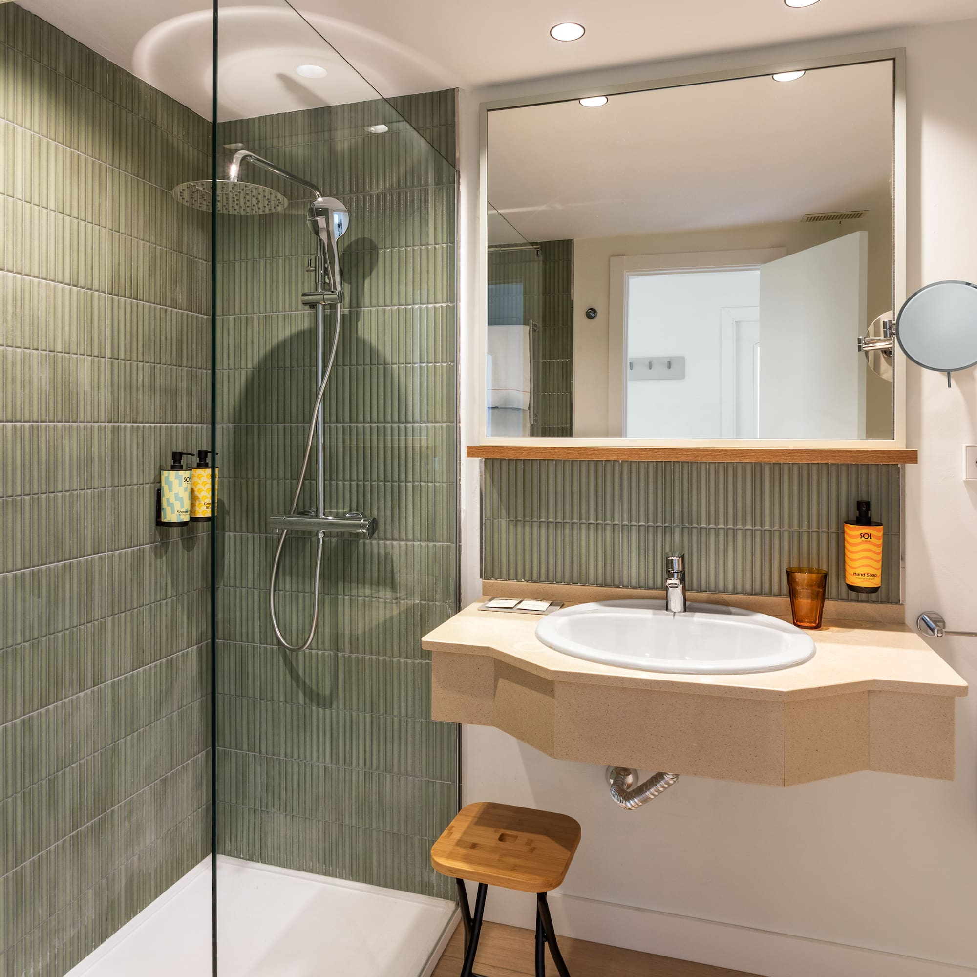 Modern hotel bathroom with green tiled shower, rain head, vanity, sink, and mirror.