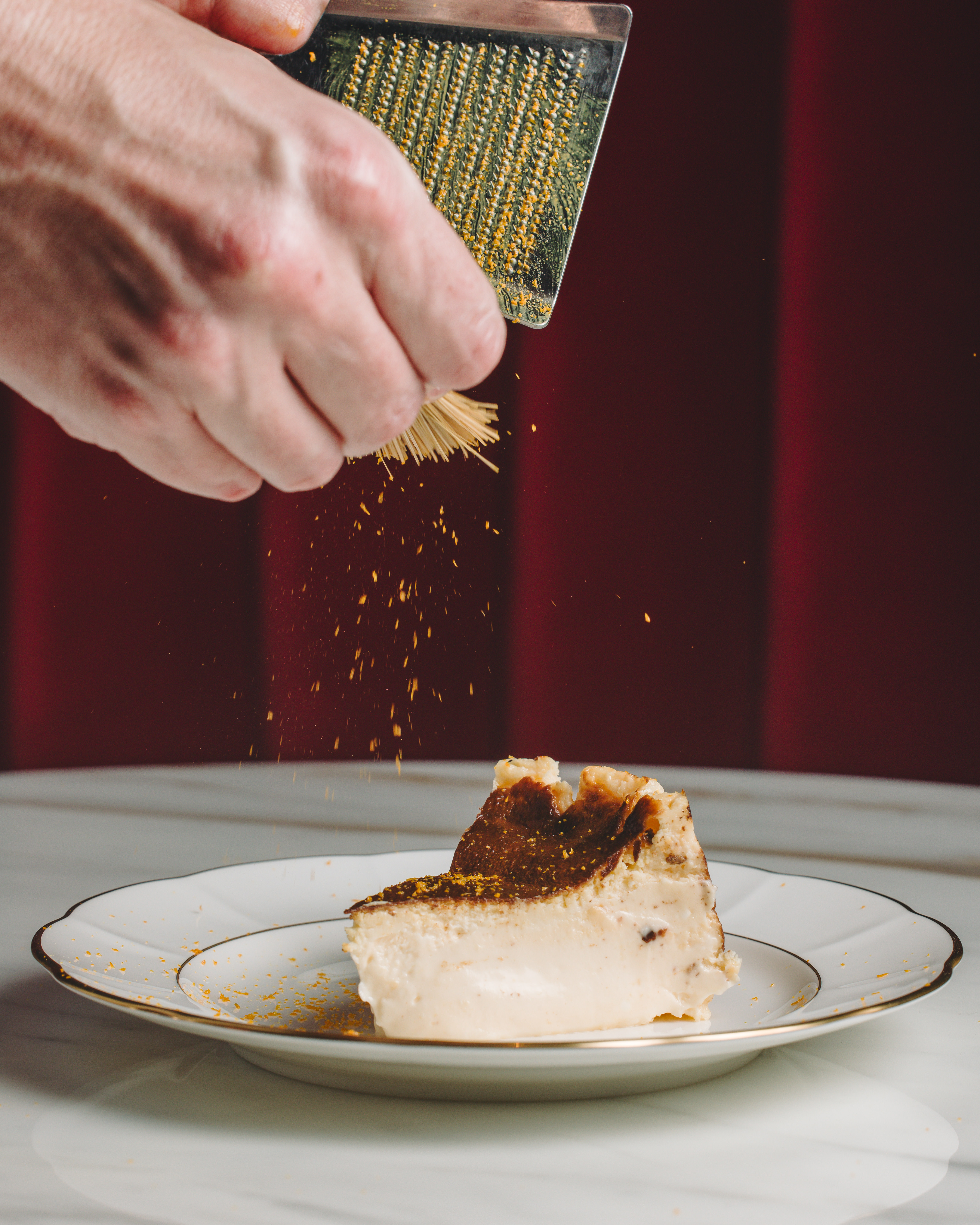 Hand grating orange zest over Basque cheesecake slice on white plate.