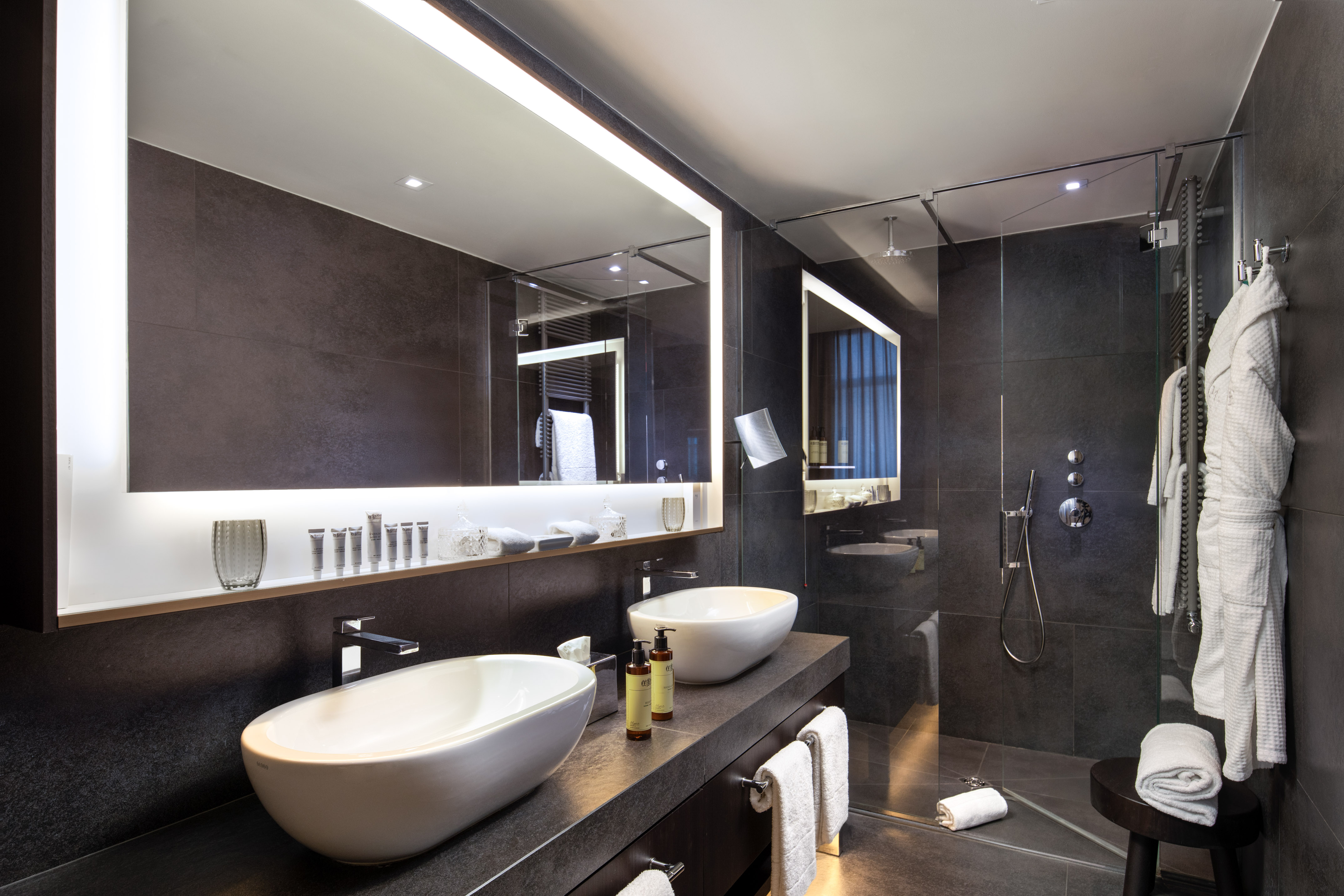 Luxury hotel bathroom with double vanity, lighted mirrors, and glass shower.