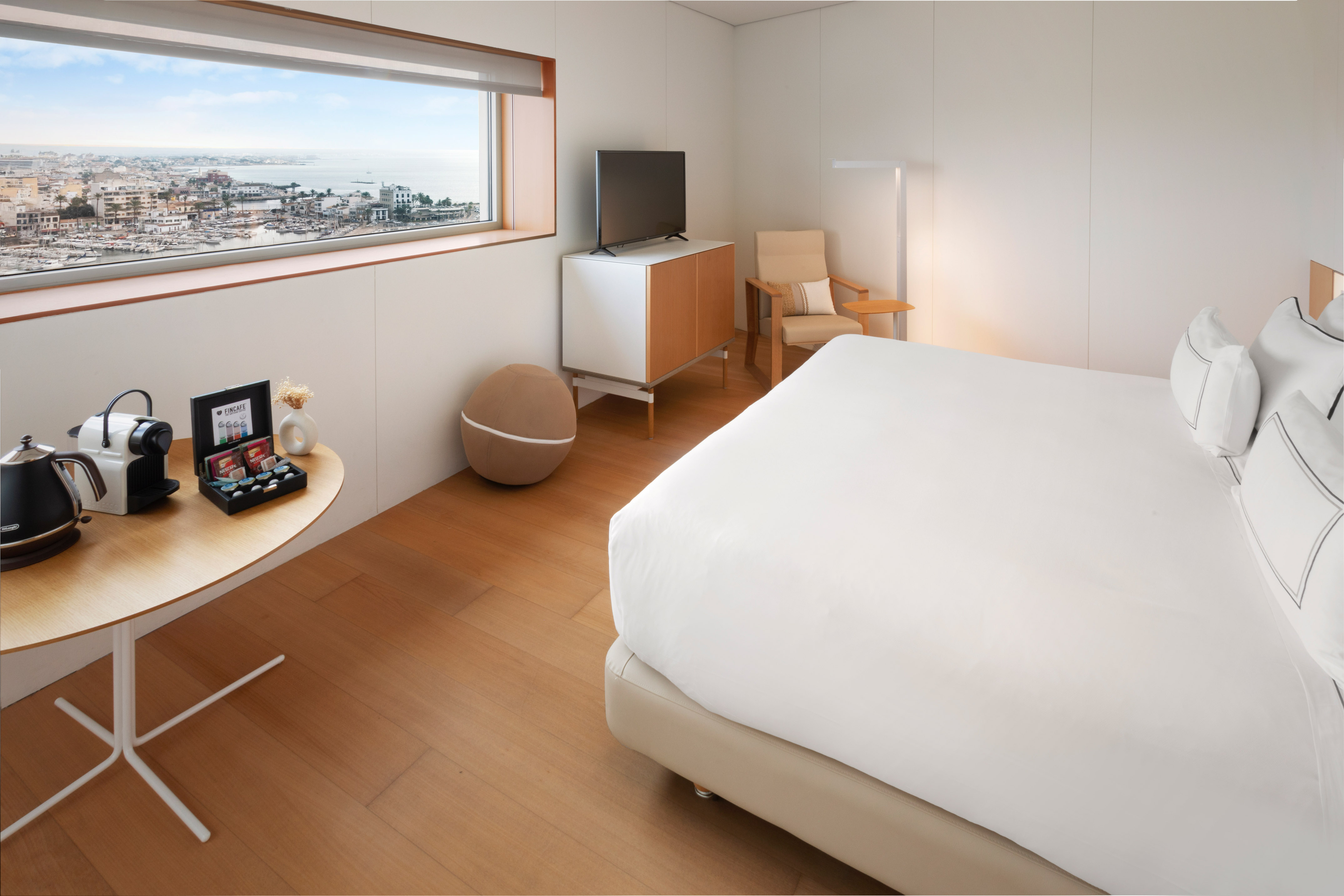 Modern hotel room with harbor view, white bed, coffee machine, and TV.