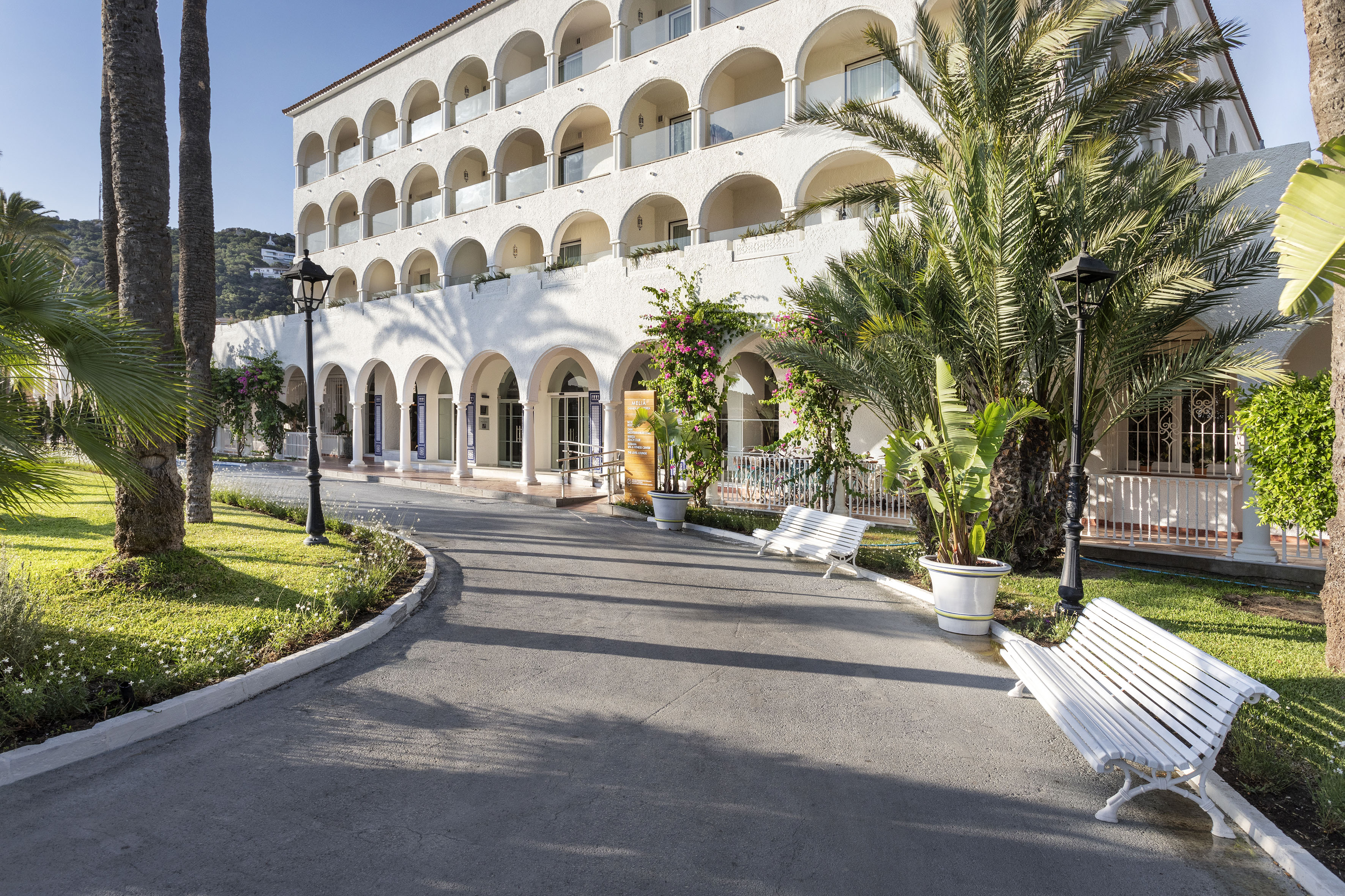 Sunny resort hotel with arched balconies, palm trees, and a paved entrance.