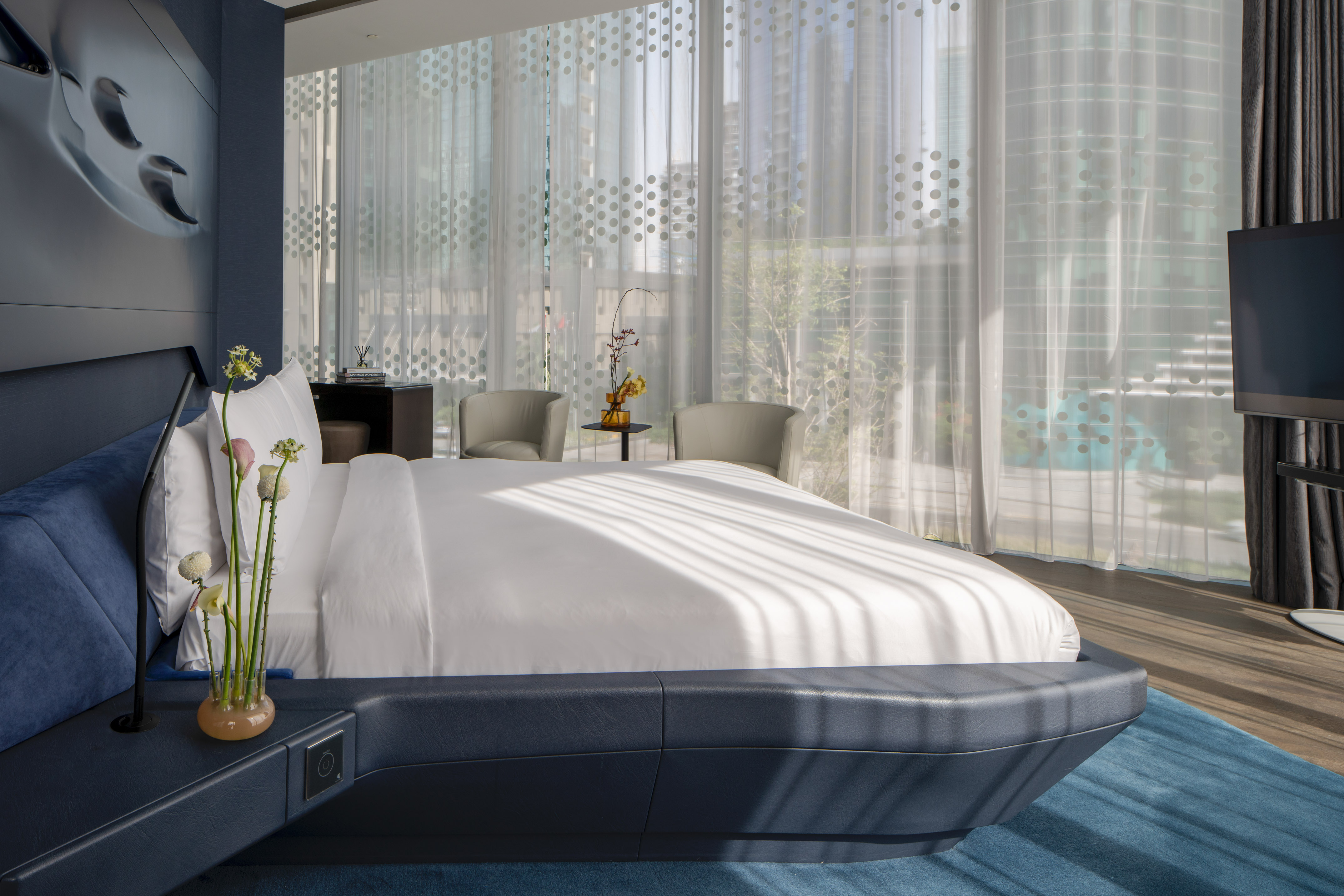 Modern hotel bedroom with a blue bed, white linens, and sheer dotted curtains.
