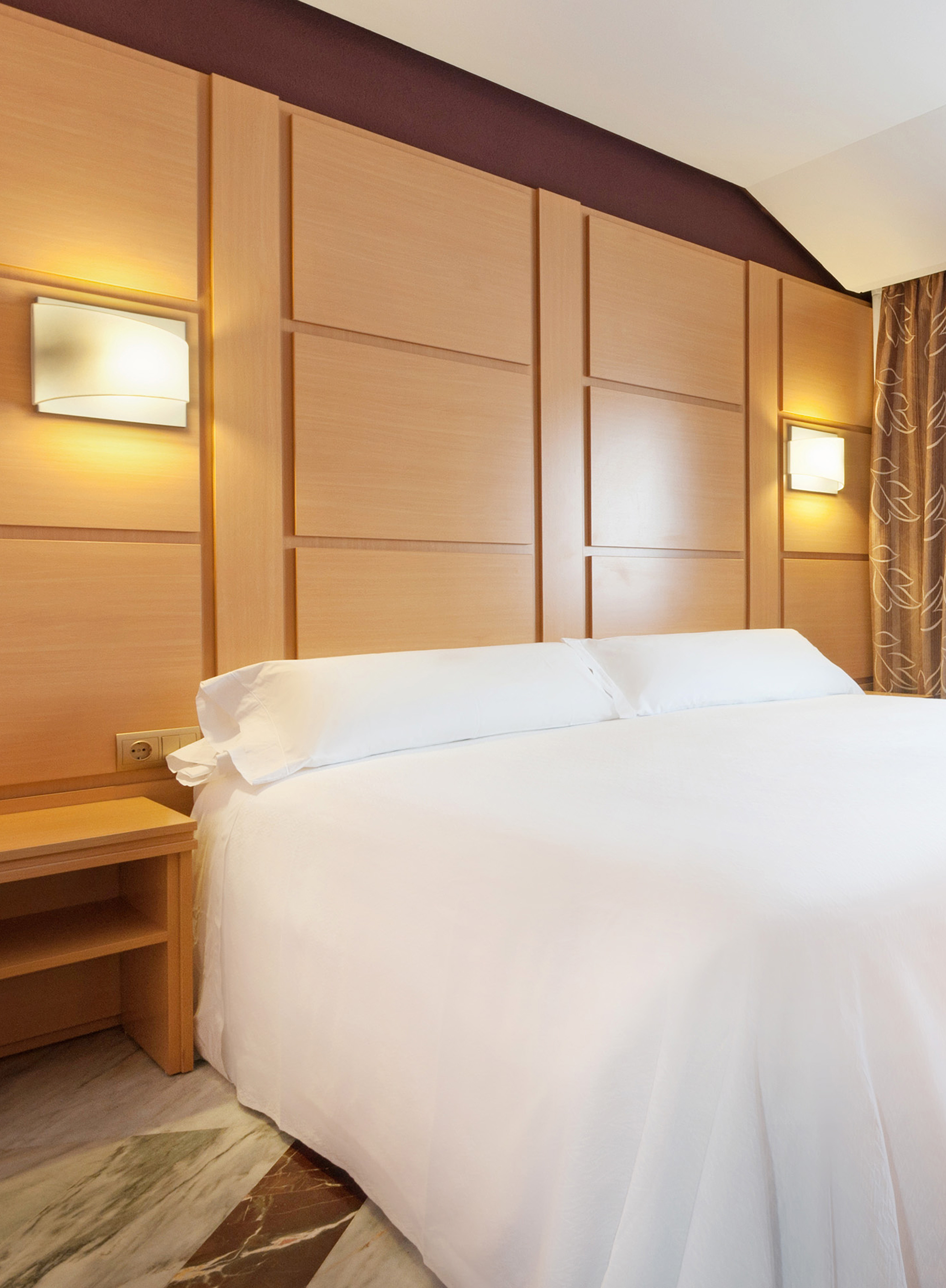Modern hotel bedroom: white bed, wooden headboard with lit sconces.