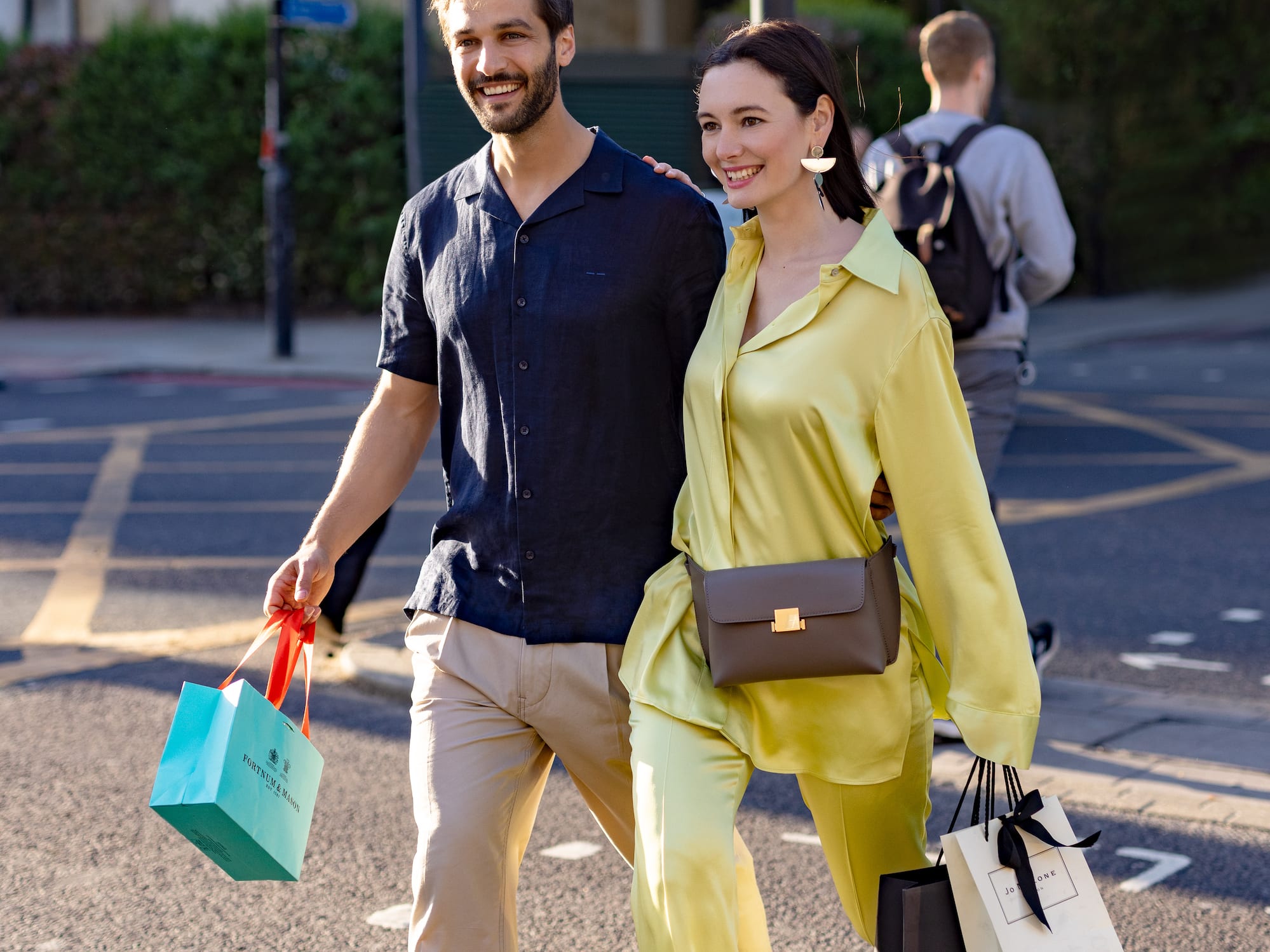 A man and a woman stroll along the street, each carrying shopping bags.
