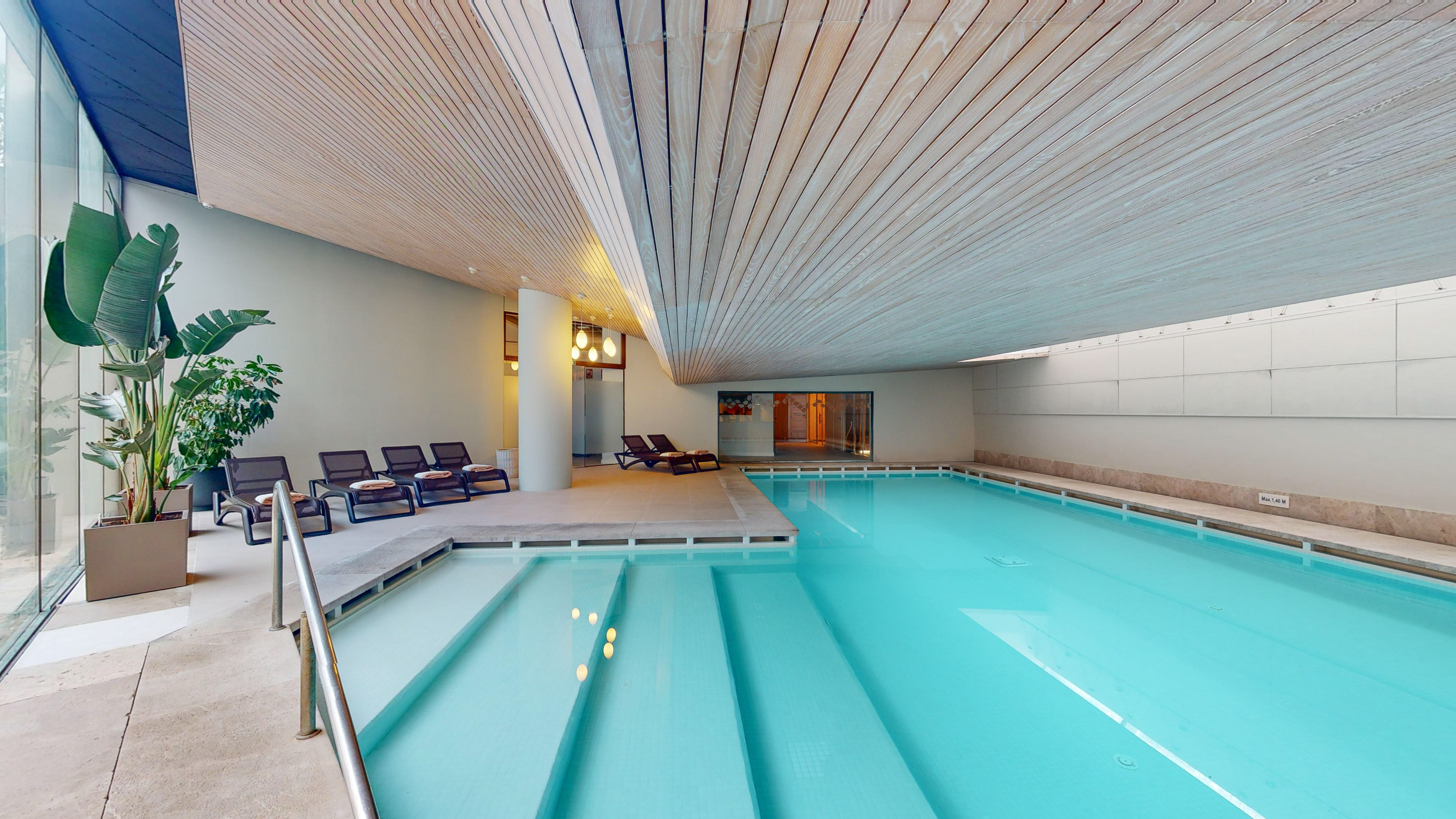 Indoor spa pool with modern wooden ceiling, lounge chairs, and plants.