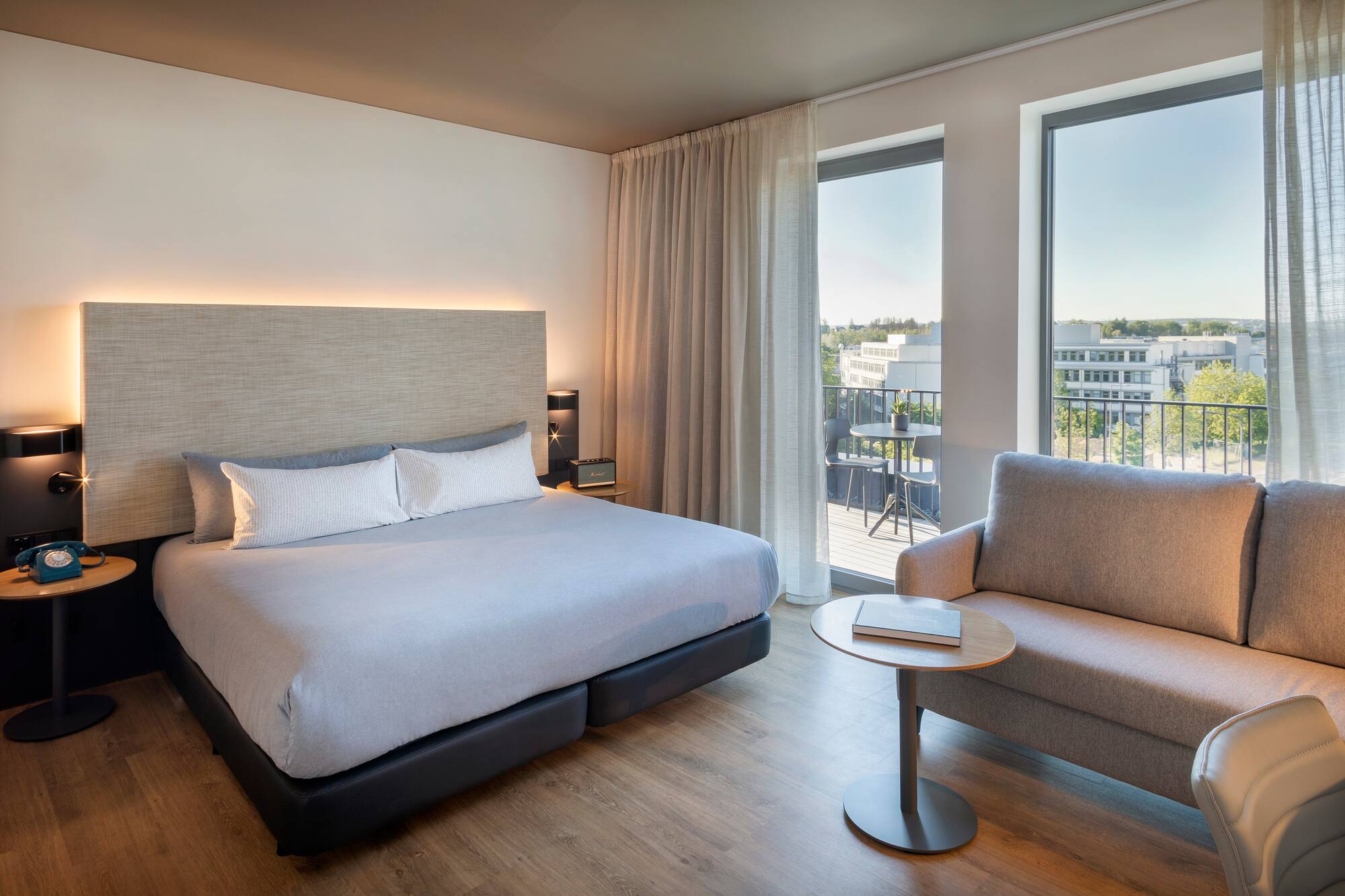 Modern hotel room with king bed, sofa, wooden floor, and city view from balcony.