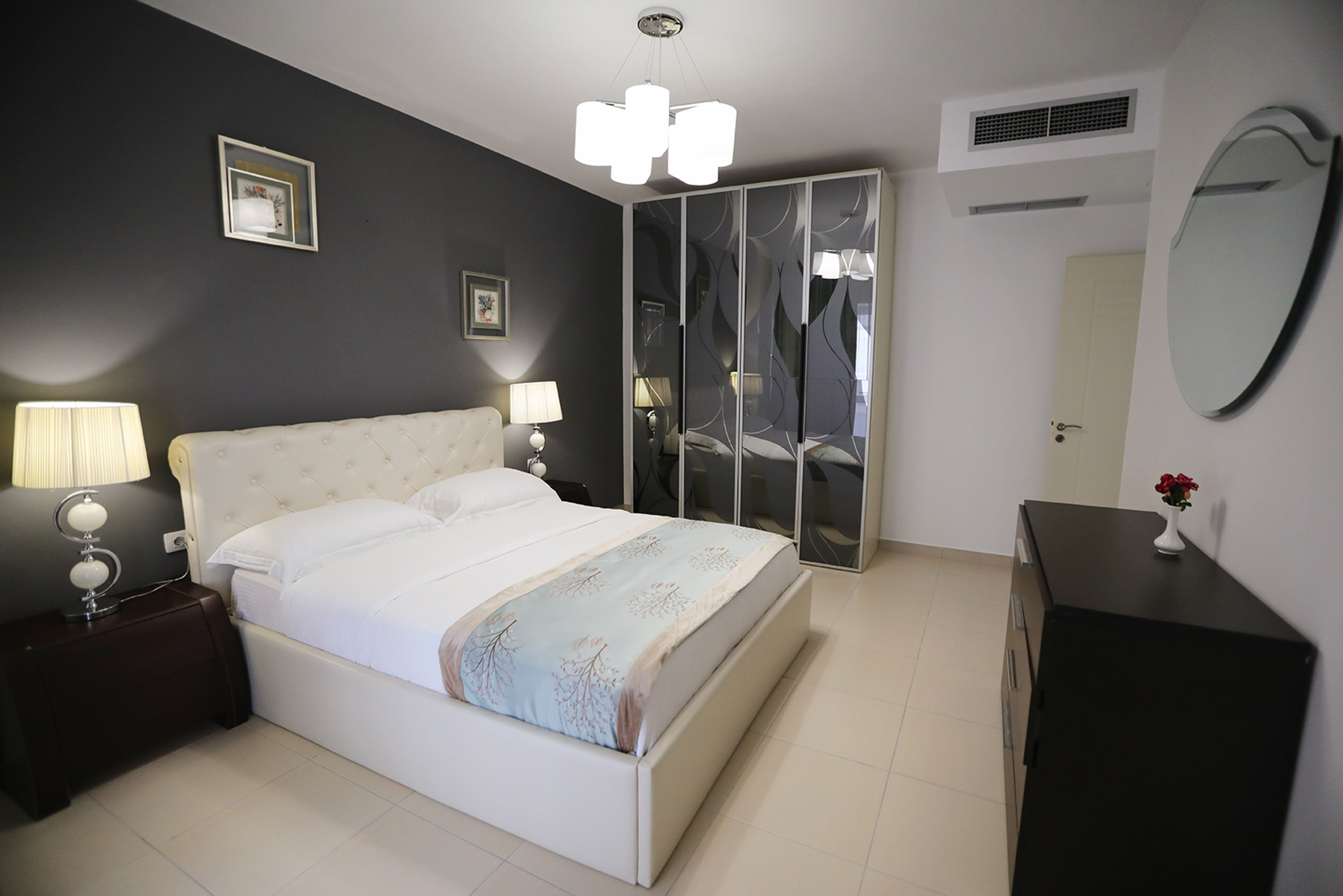 Modern bedroom with white bed, tufted headboard, dark grey wall, and mirrored wardrobe.