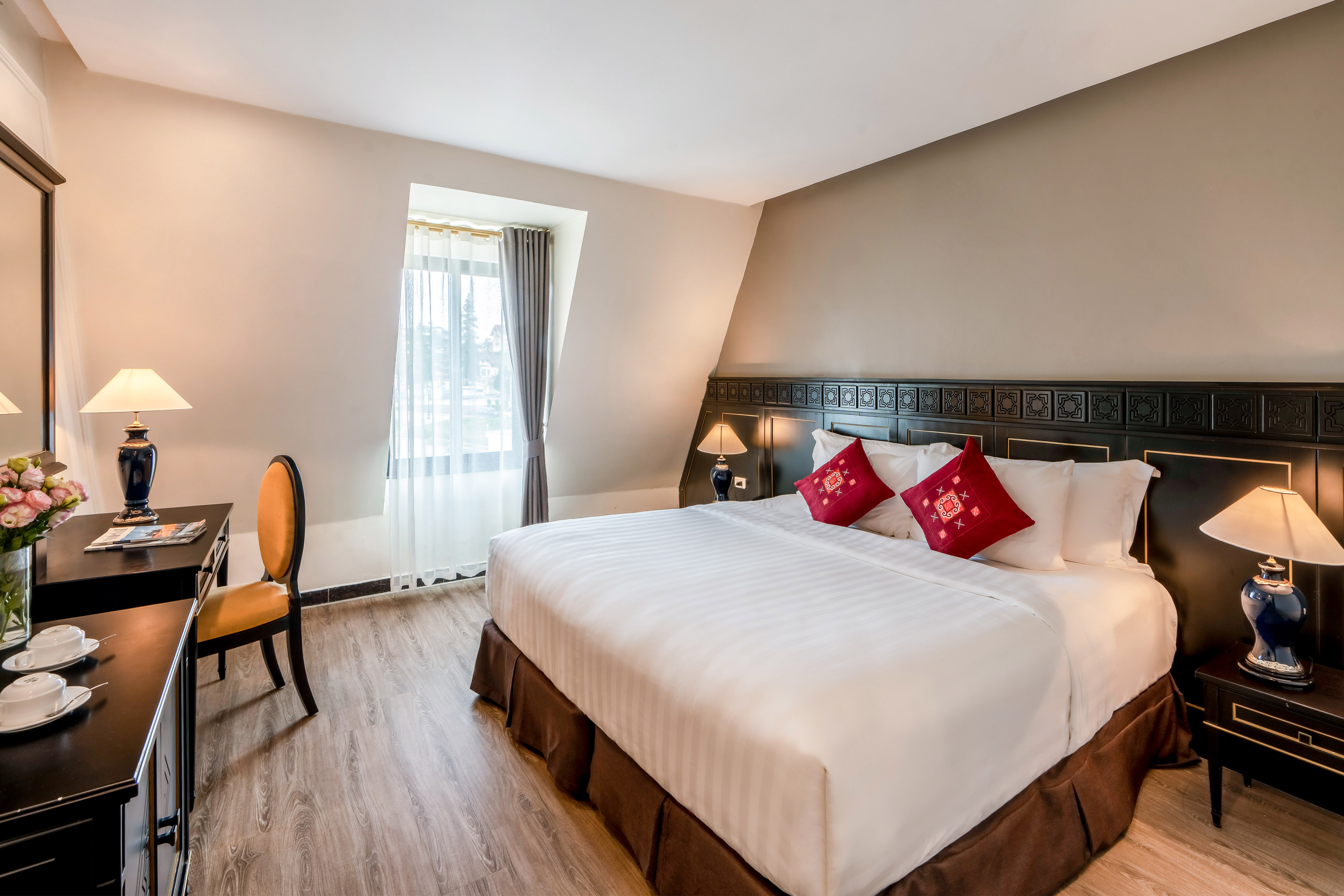 Modern hotel bedroom with king bed, red pillows, ornate headboard, and desk.