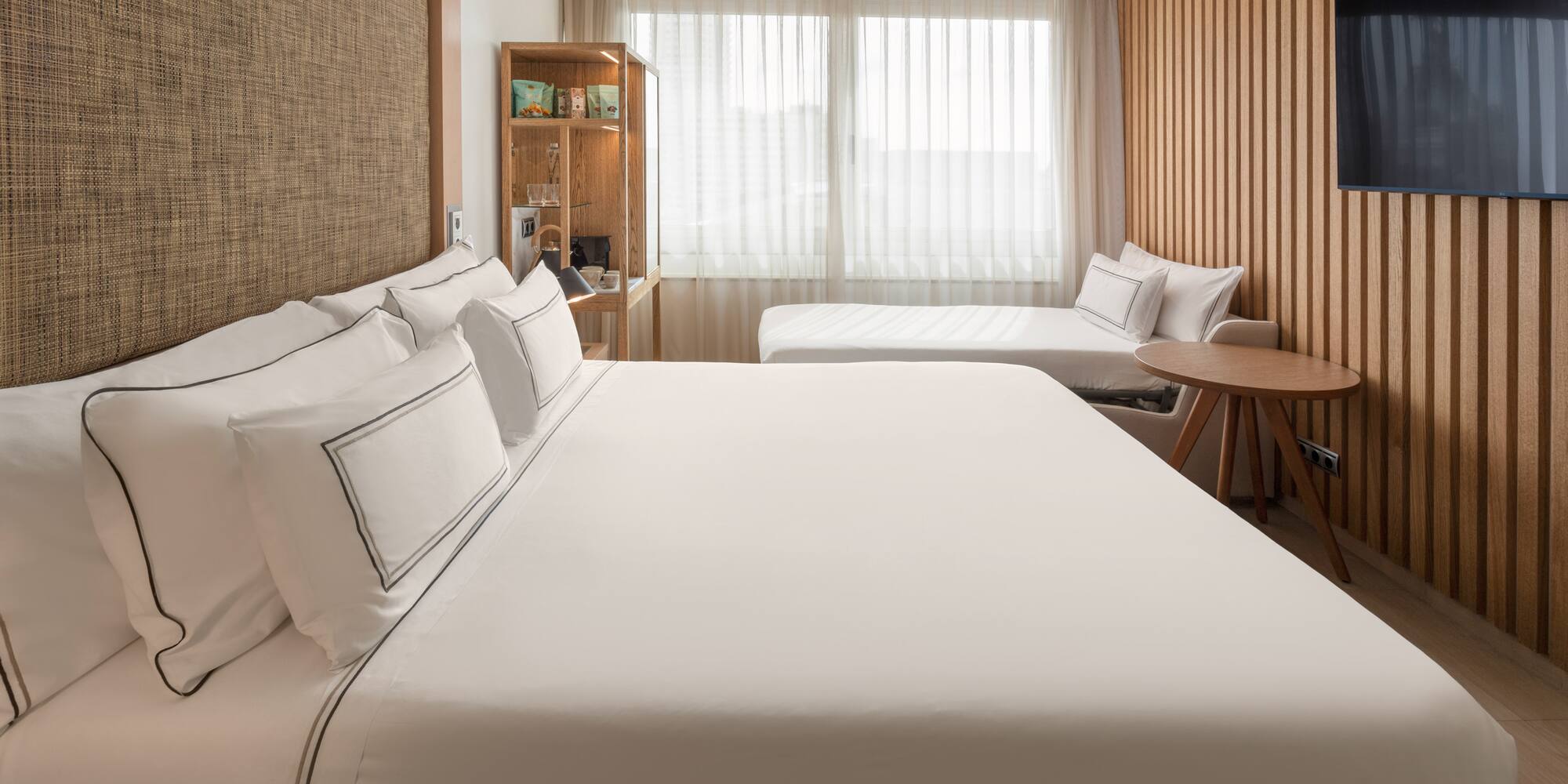 Modern hotel room with large bed, daybed, wood paneling, and bright window.