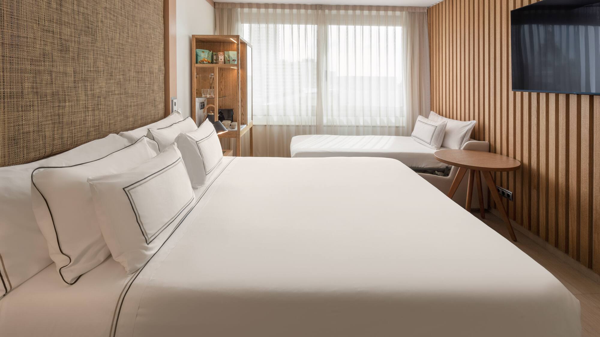 Modern hotel room with large bed, daybed, wood paneling, and bright window.