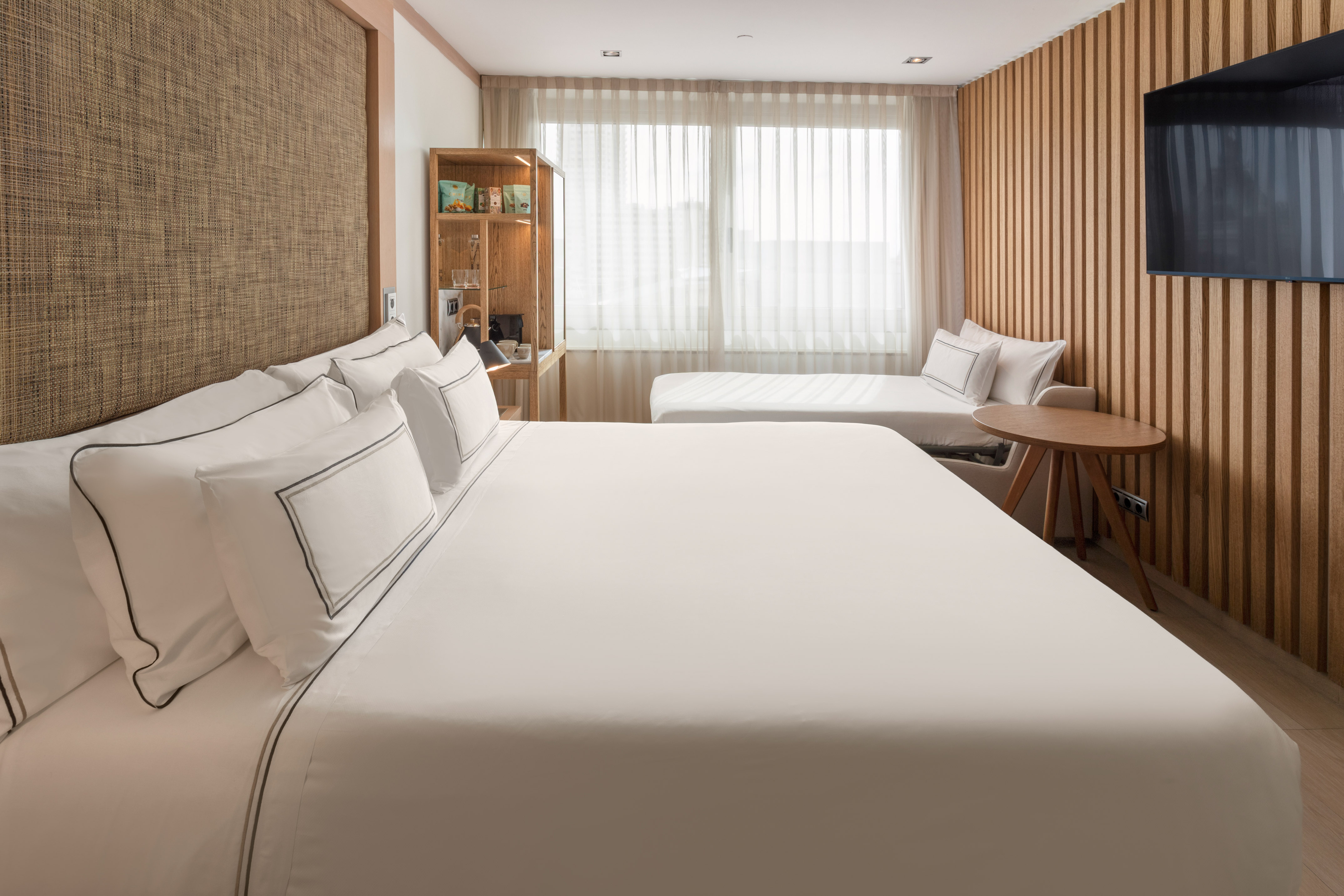 Bright modern hotel room with bed, daybed, wooden wall, and wall-mounted TV.