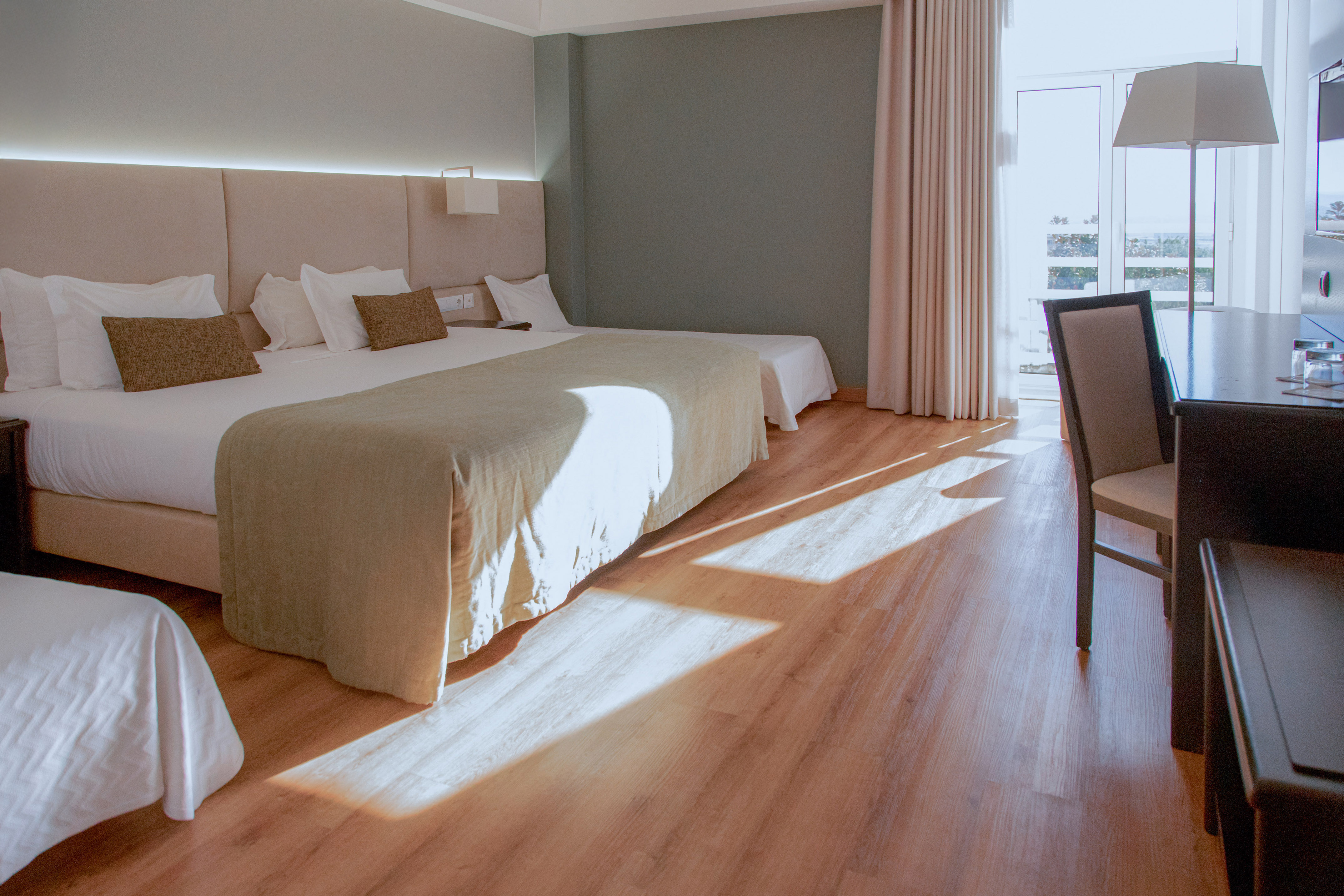 Bright hotel room with large bed, extra bed, desk, and wood floor.