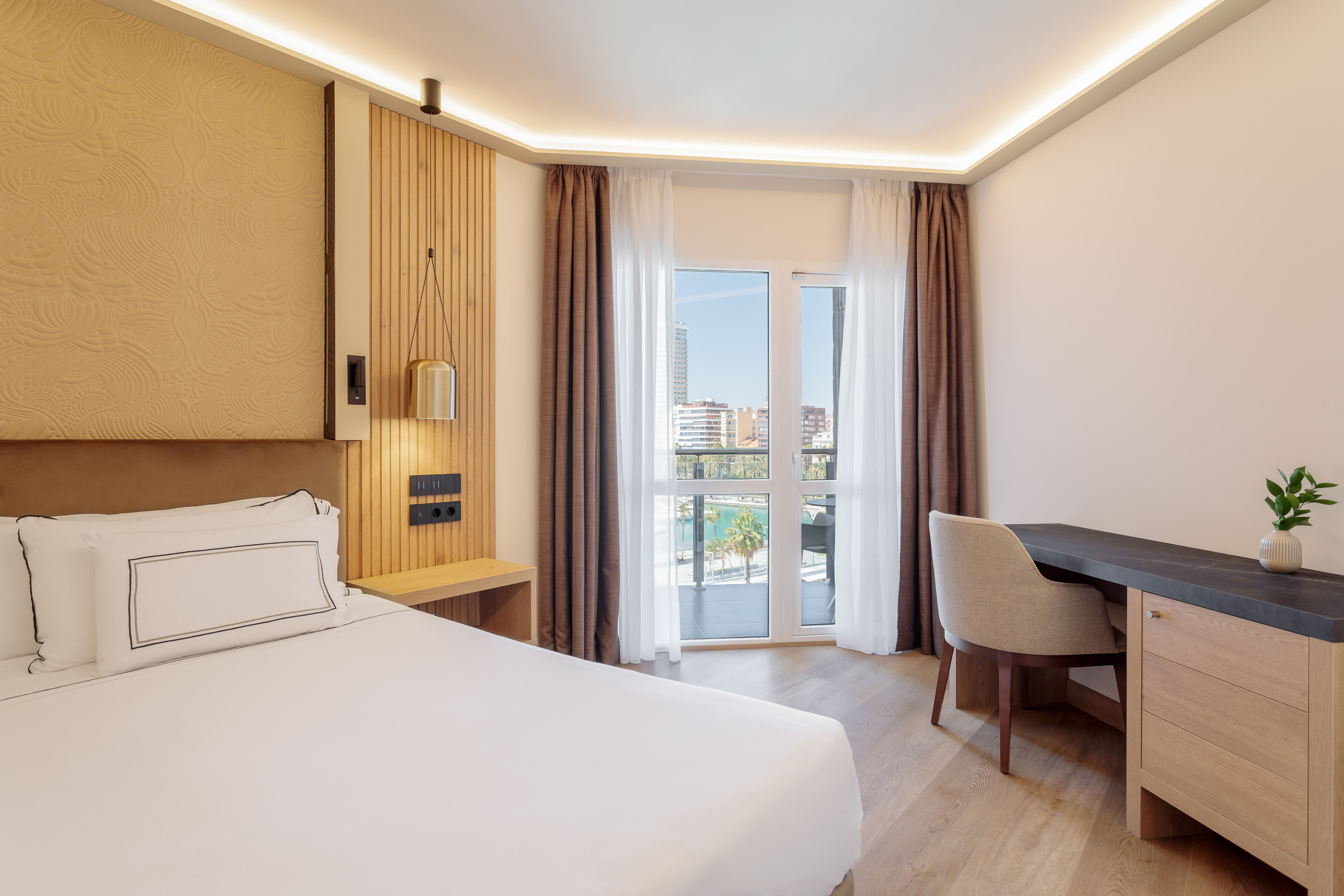 Modern hotel bedroom with white bed, desk, and waterfront city view.