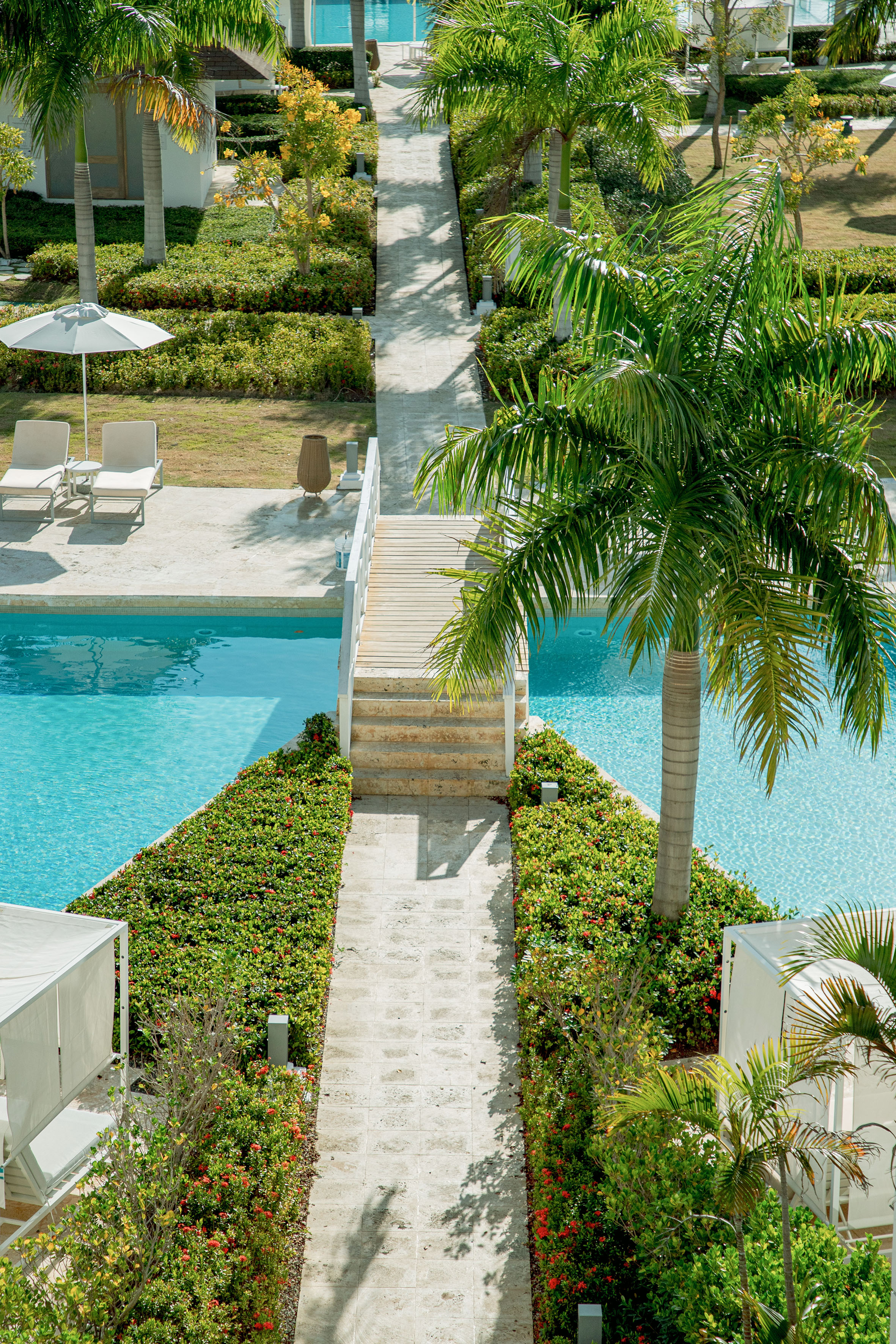 Tropical resort pool with palm trees, stone path, and wooden bridge over aqua water.