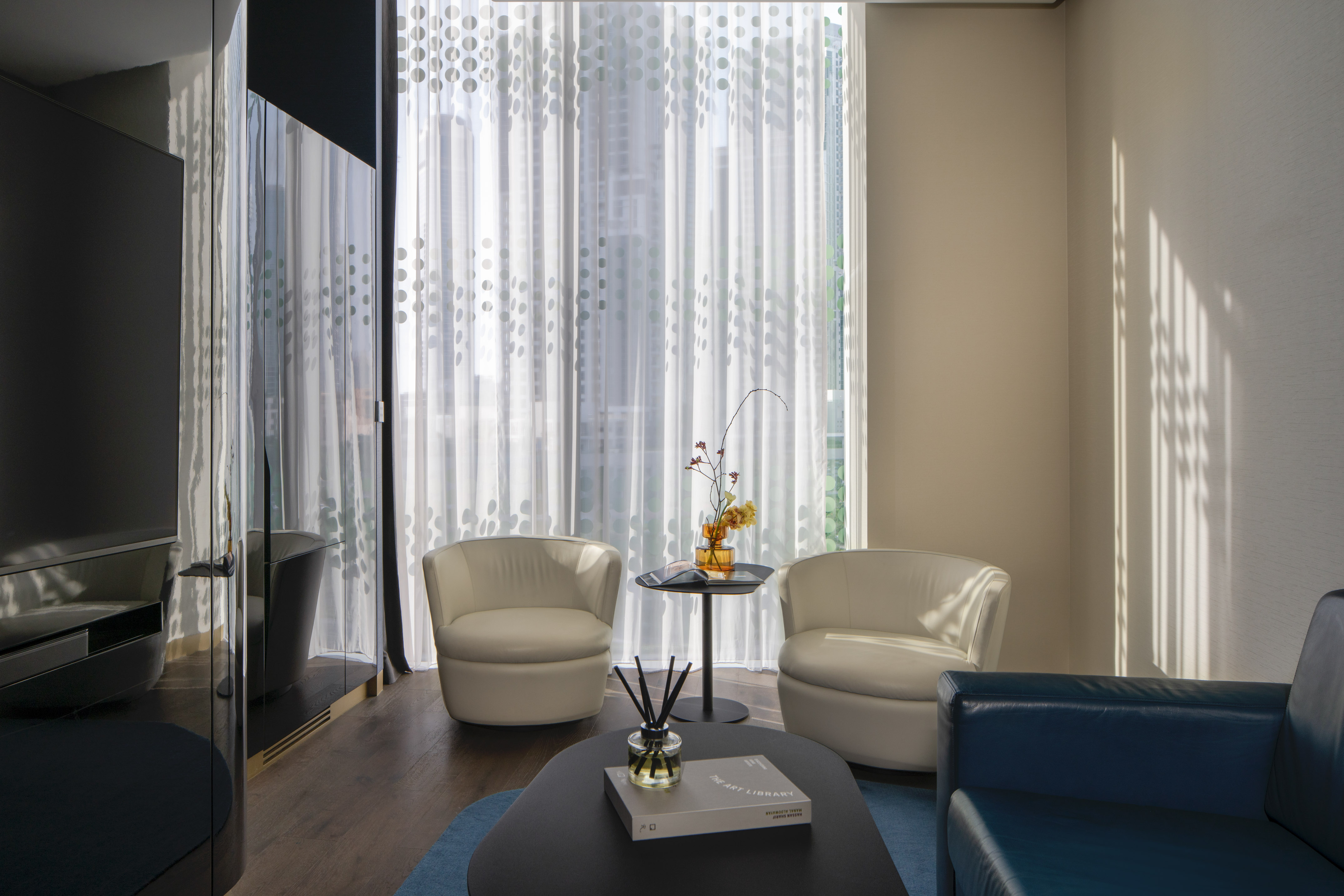 Modern lounge with white armchairs, blue sofa, patterned curtains, sunlit window.