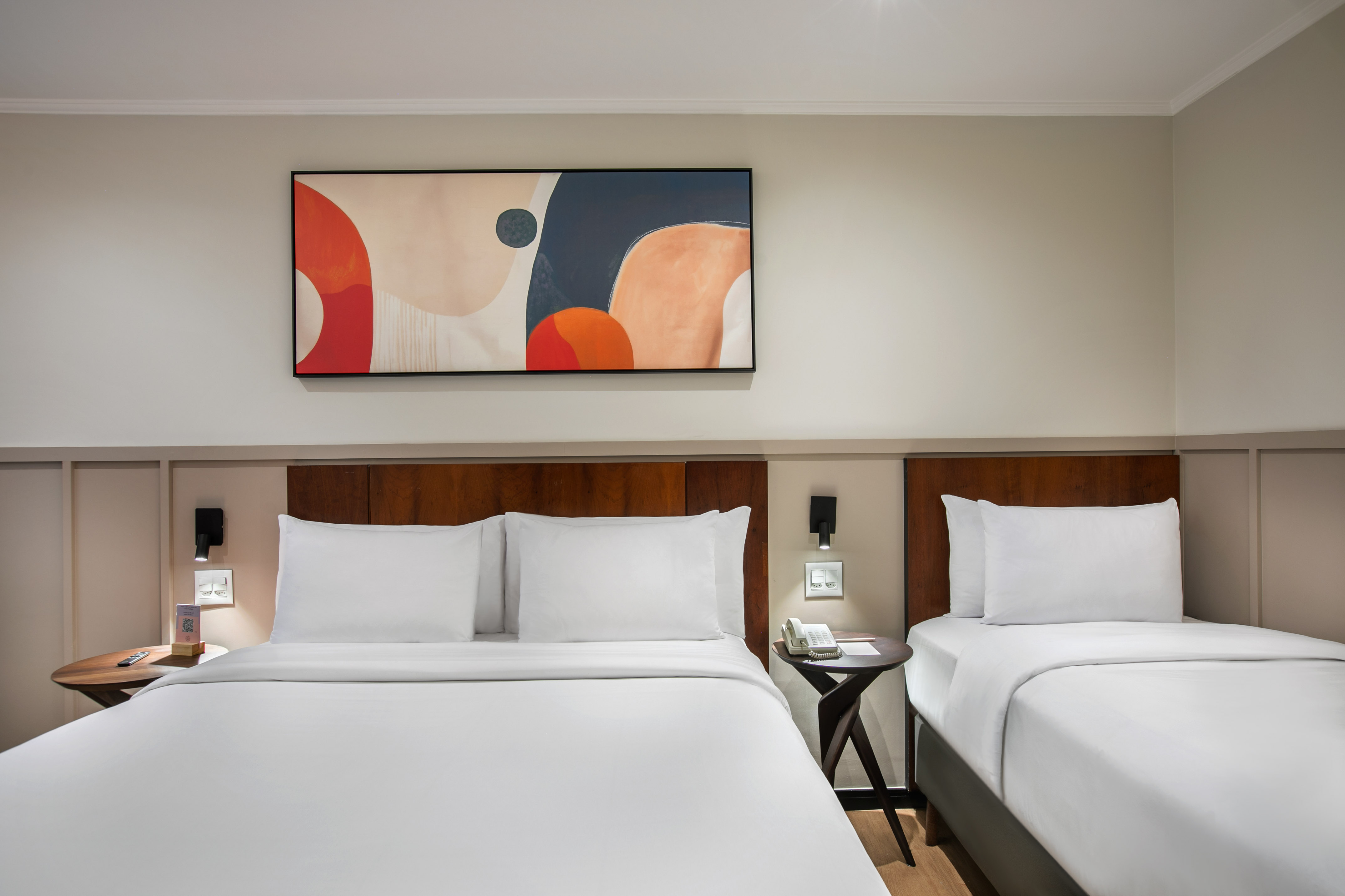 Modern hotel room with two beds, white linens, wooden headboards, abstract art.