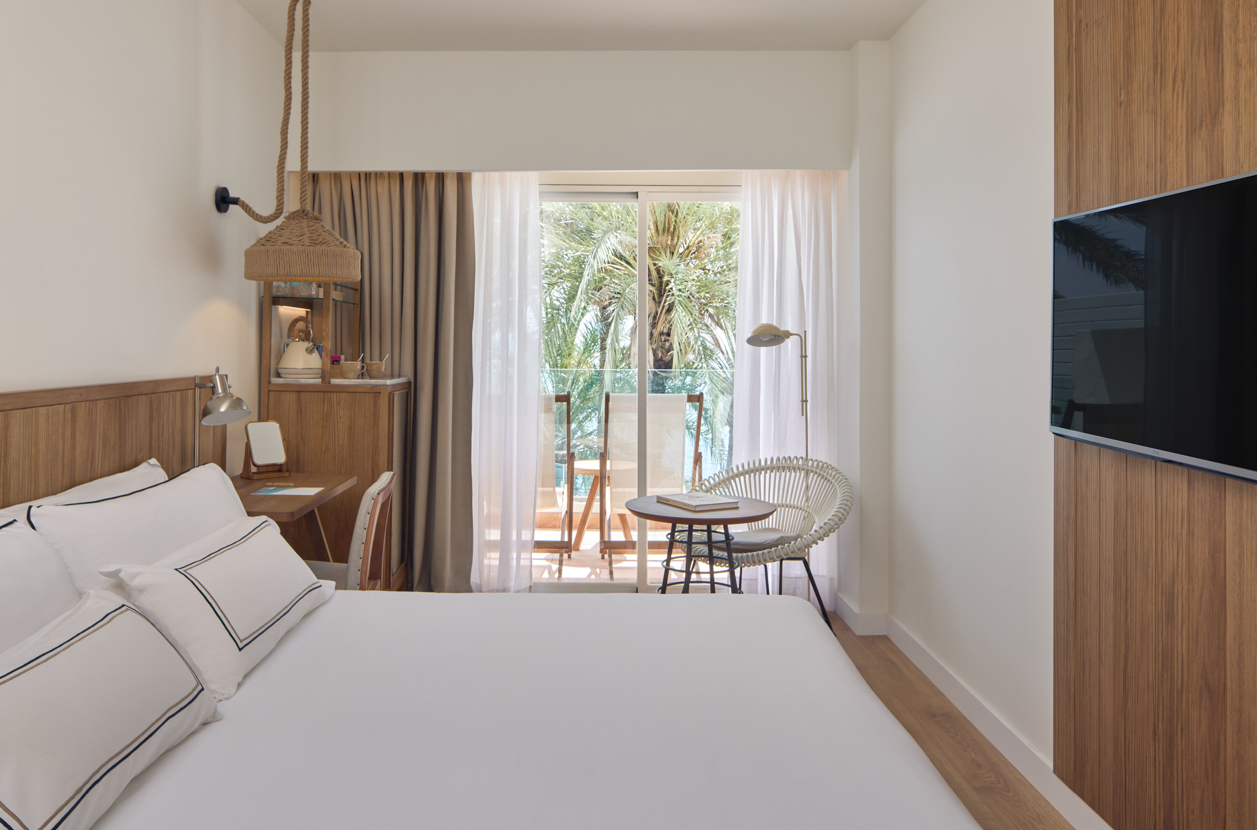 Modern hotel room with white bed, desk, and balcony overlooking tropical palm trees.