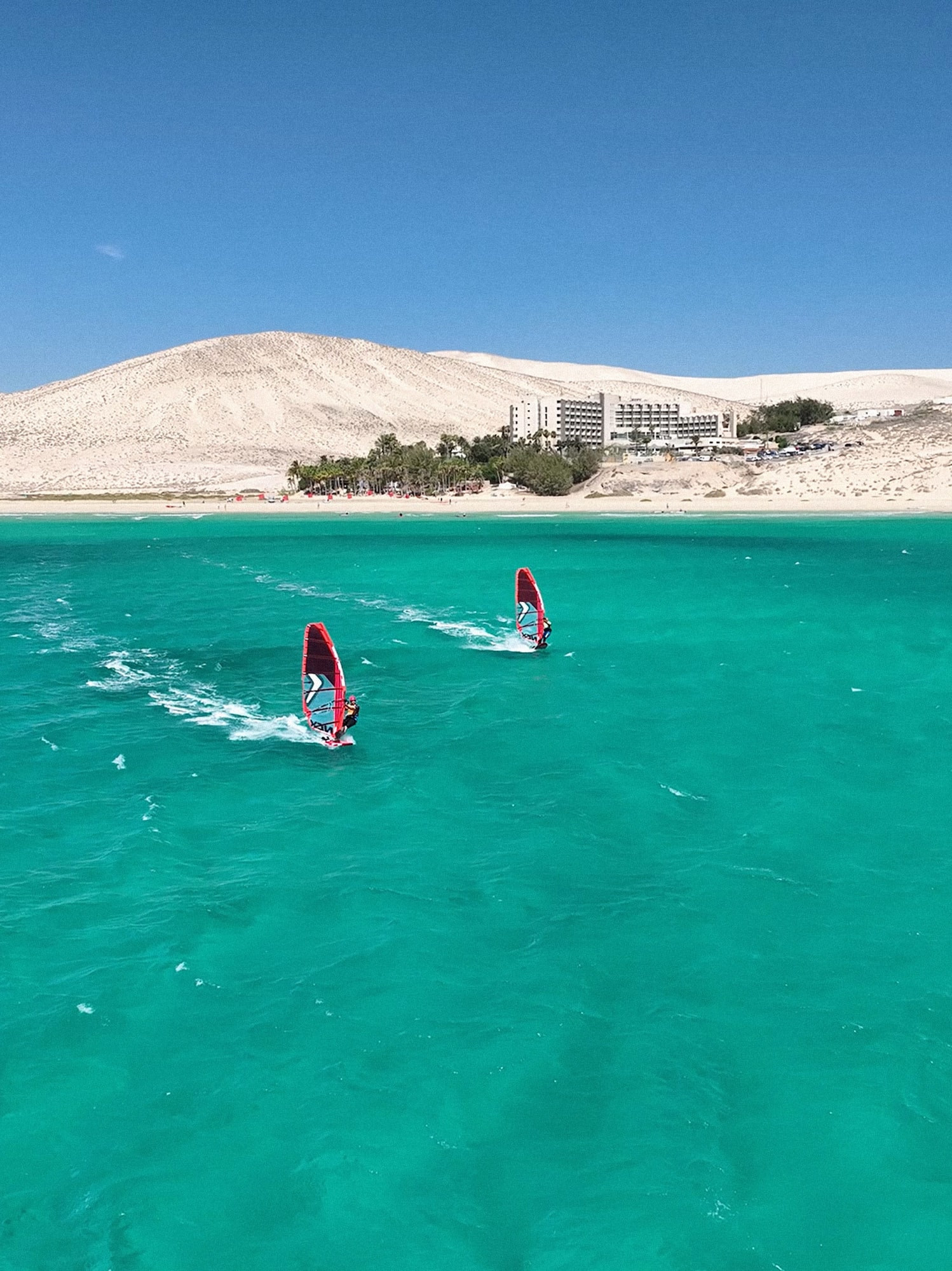 Windsurfers on turquoise sea with sandy beach, sand dunes, and resort hotel.