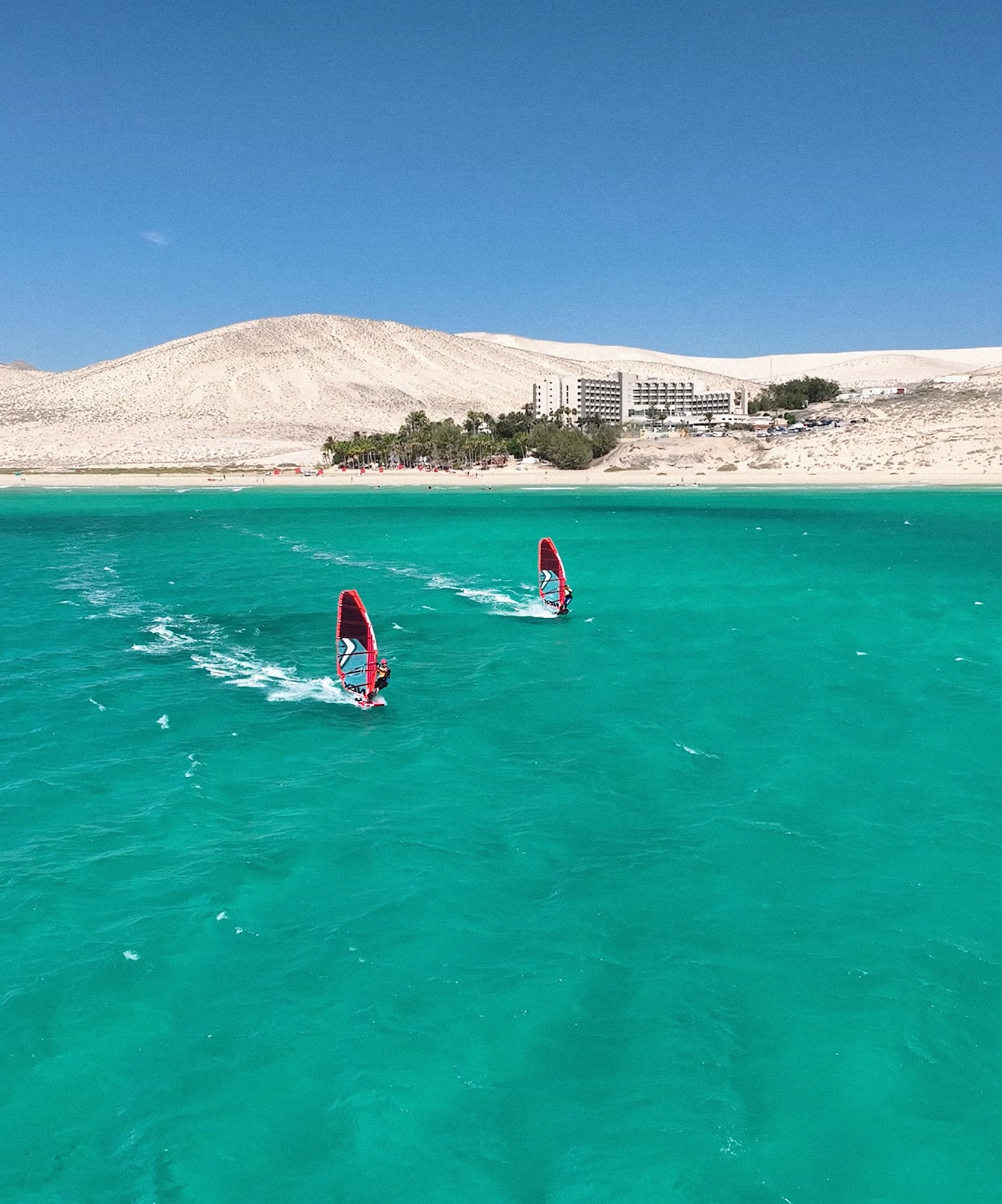 Windsurfistas en mar turquesa, dunas y hotel costero. Fuerteventura.