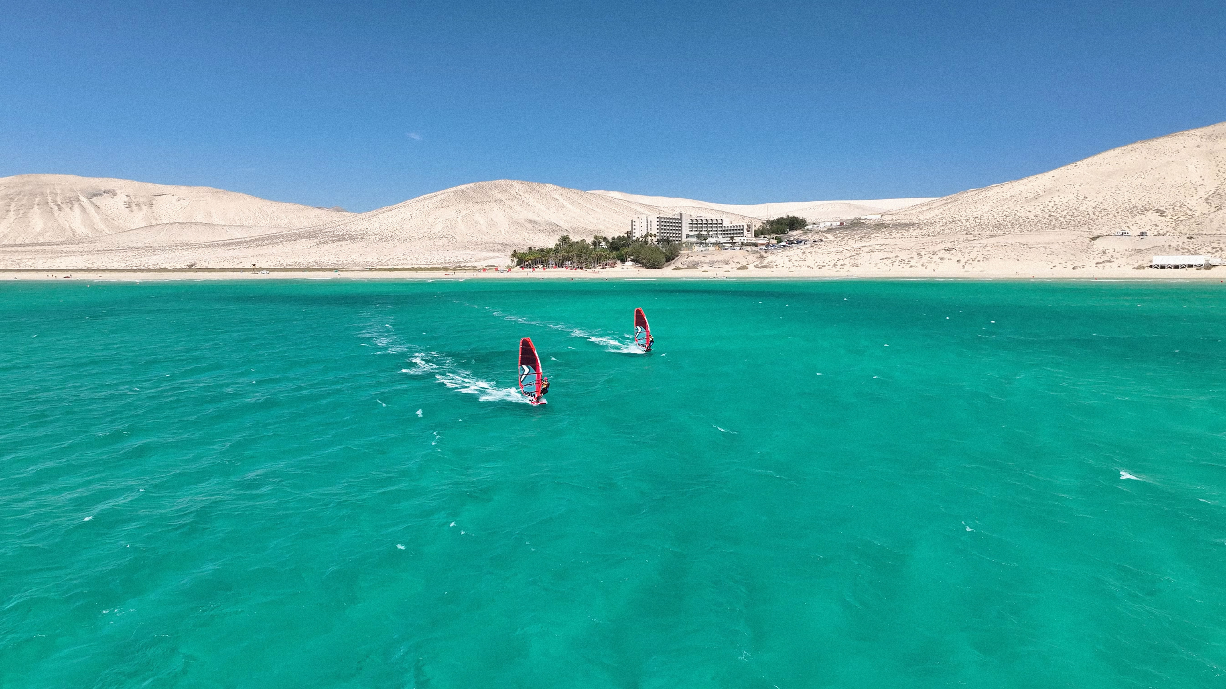 Windsurfistas en mar turquesa, dunas y hotel costero. Fuerteventura.