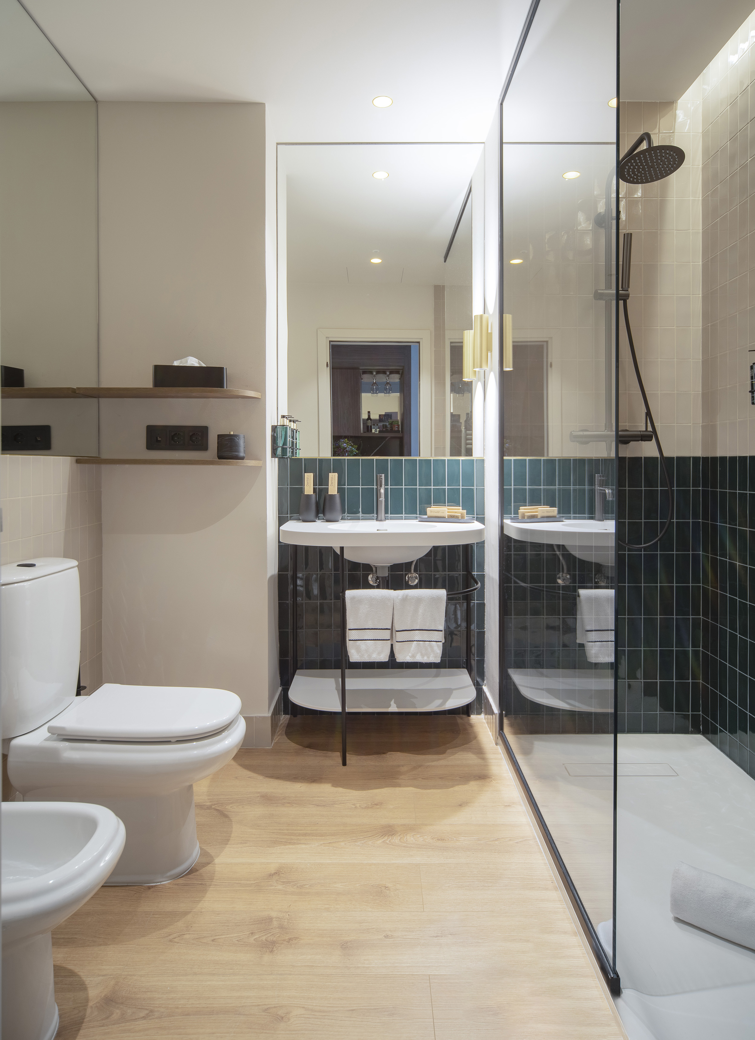 Modern bathroom with toilet, bidet, vanity, glass shower, and teal tiled walls.