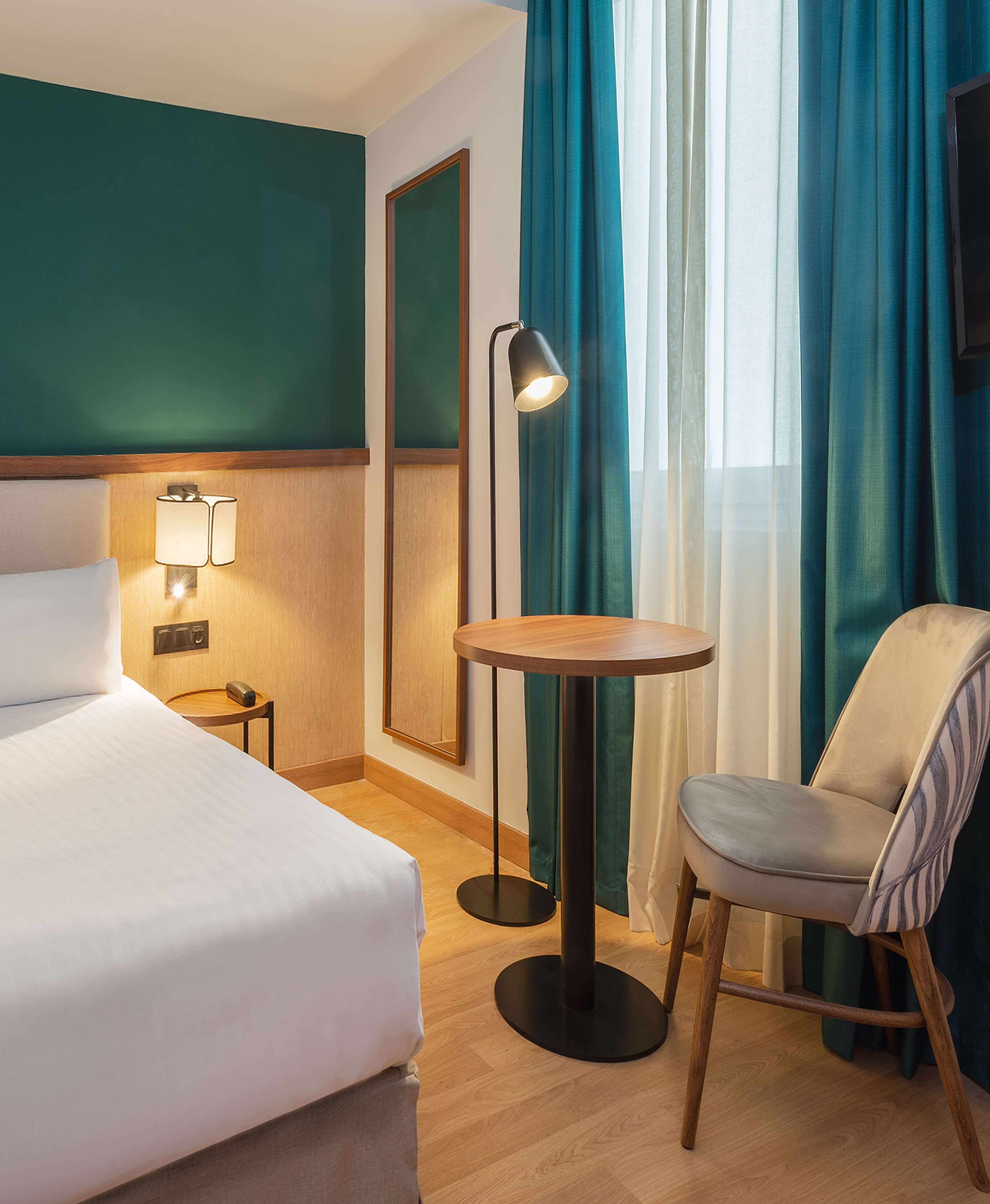 Modern hotel room with white bed, teal curtains, wooden table, and lamp.