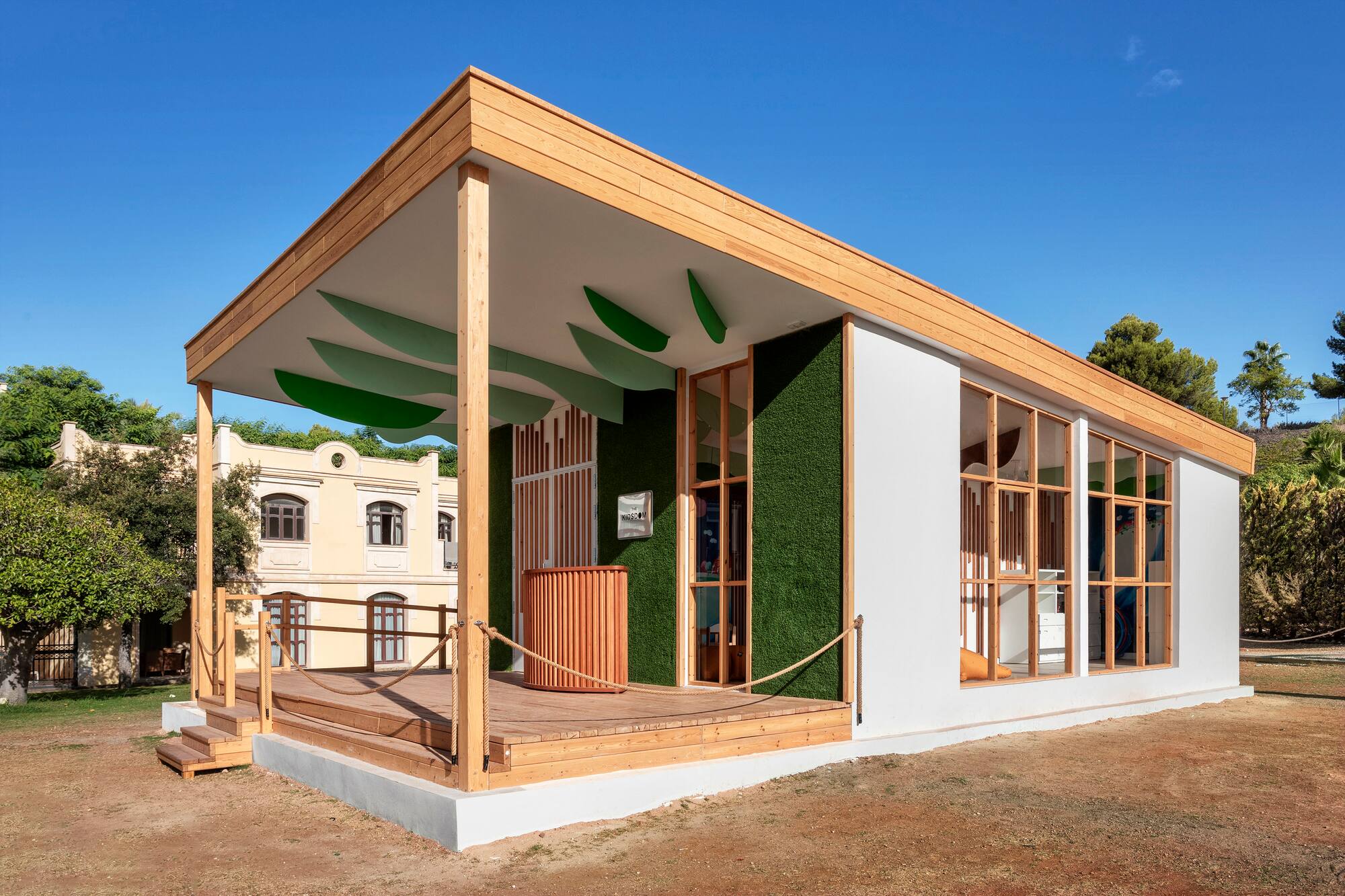 Modular wooden building with outdoor deck, green turf wall, and leaf ceiling.