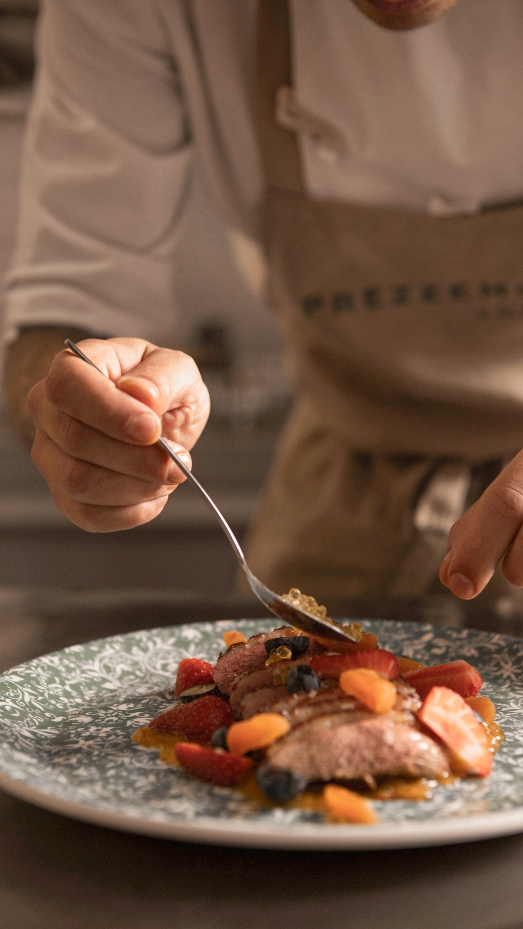 Chef plating gourmet duck breast with berries and caviar on a patterned plate.