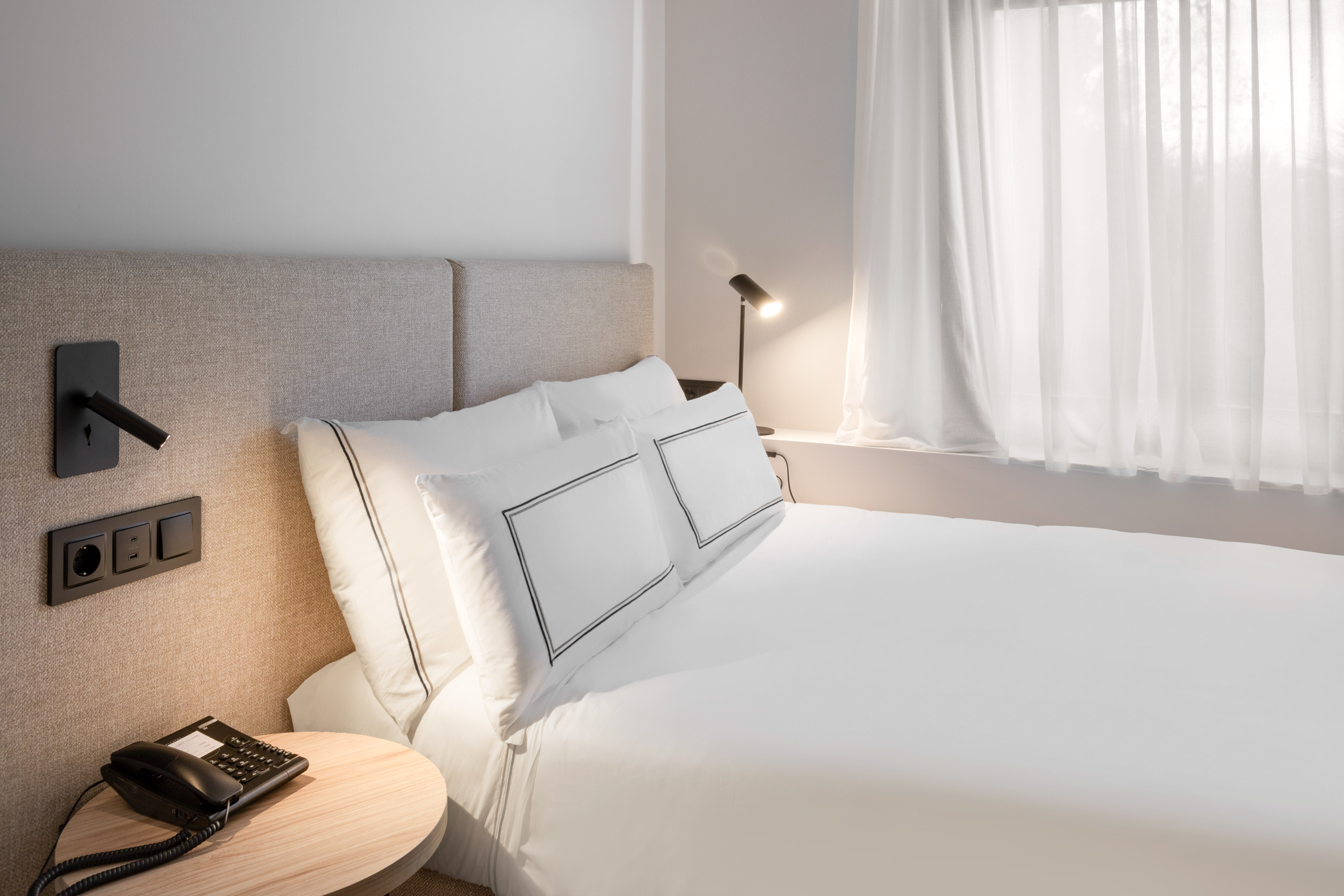 Minimalist hotel bedroom with white bed, grey headboard, and bedside lamps.