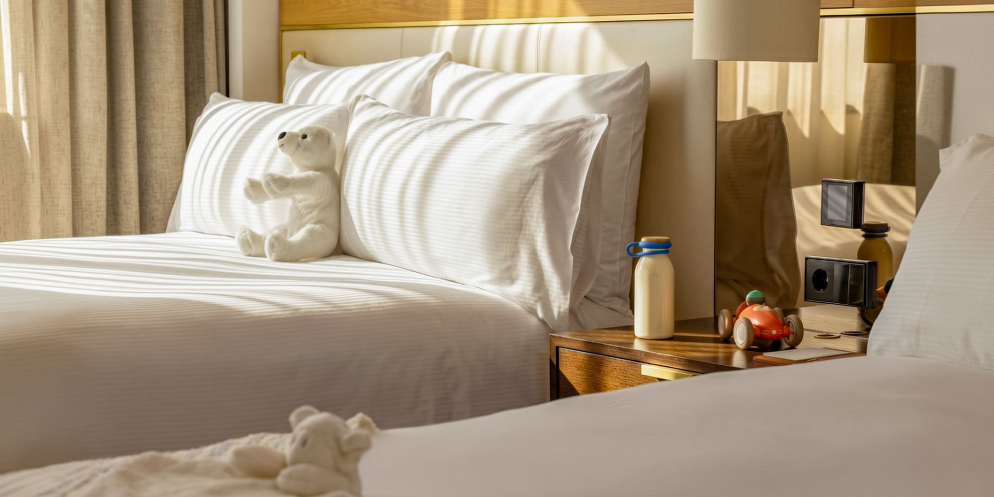 Family hotel room with twin beds, white bedding, teddy bear, and toy car.