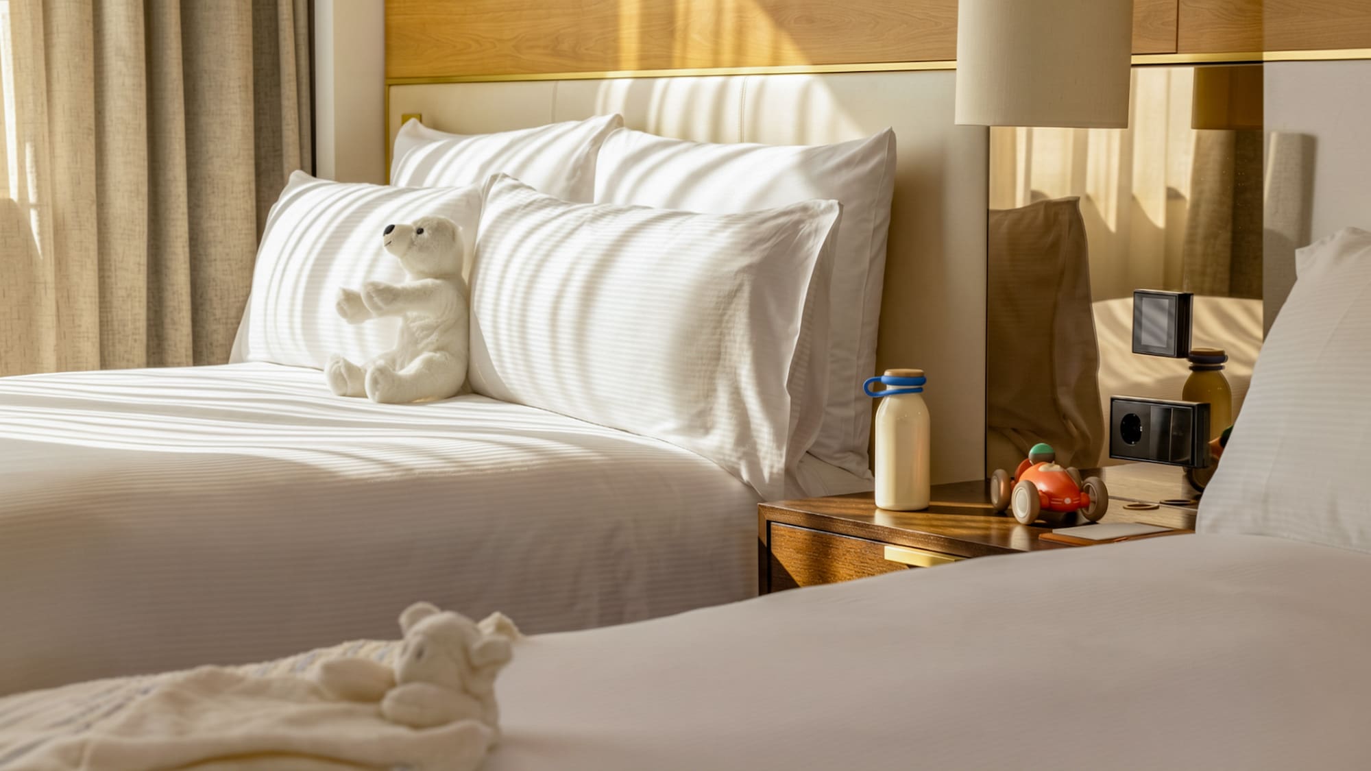 Family hotel room with twin beds, white bedding, teddy bear, and toy car.