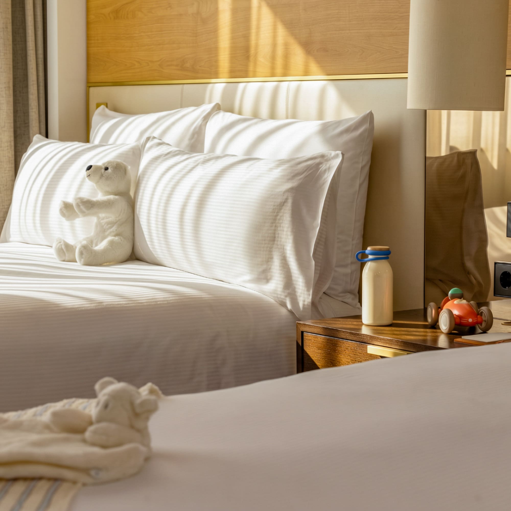 Family hotel room with twin beds, white bedding, teddy bear, and toy car.